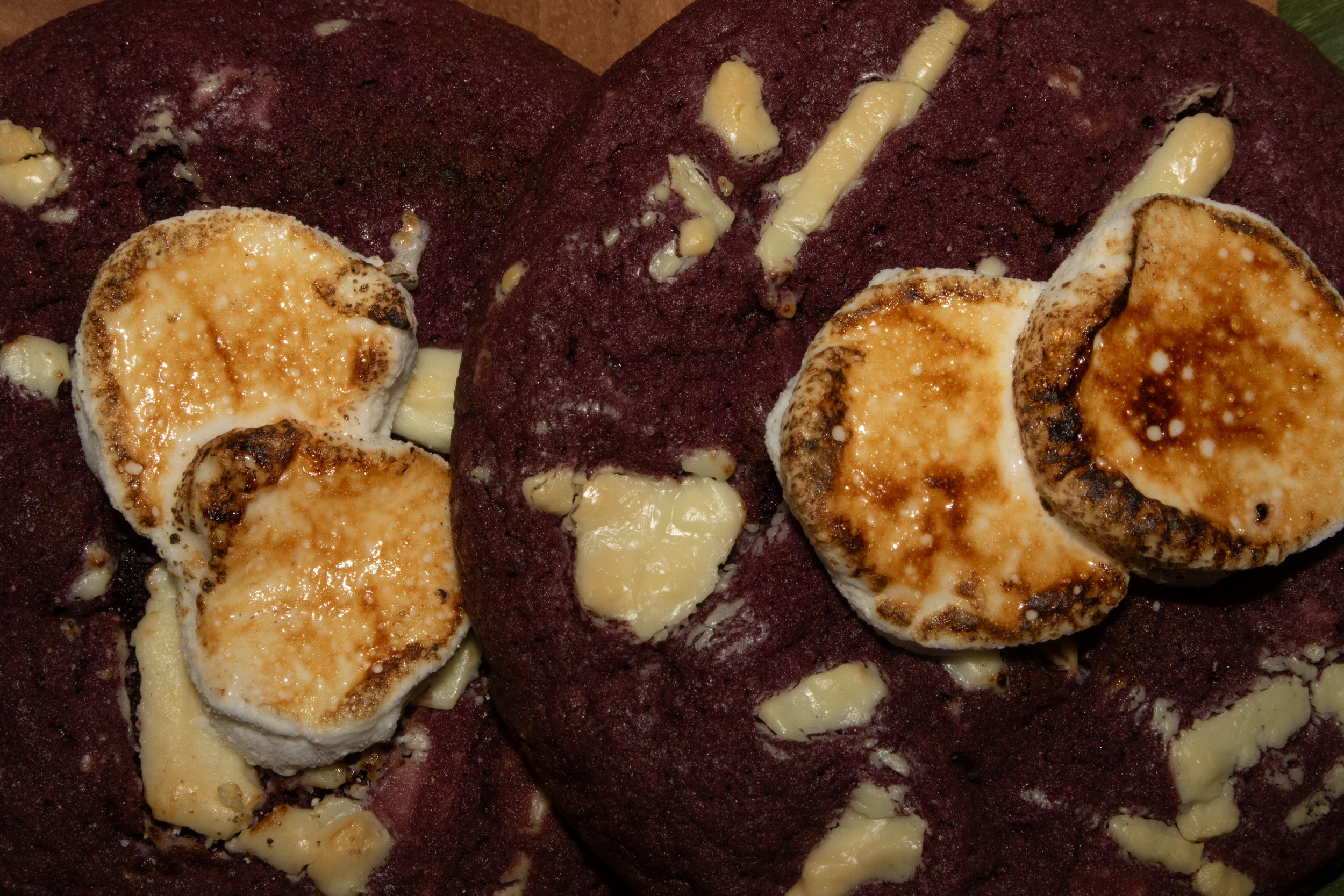 Close-up image of toasted marshmallows with a browned surface, some melted cheese, and a dark purple cake with white streaks and bits of melted cheese.