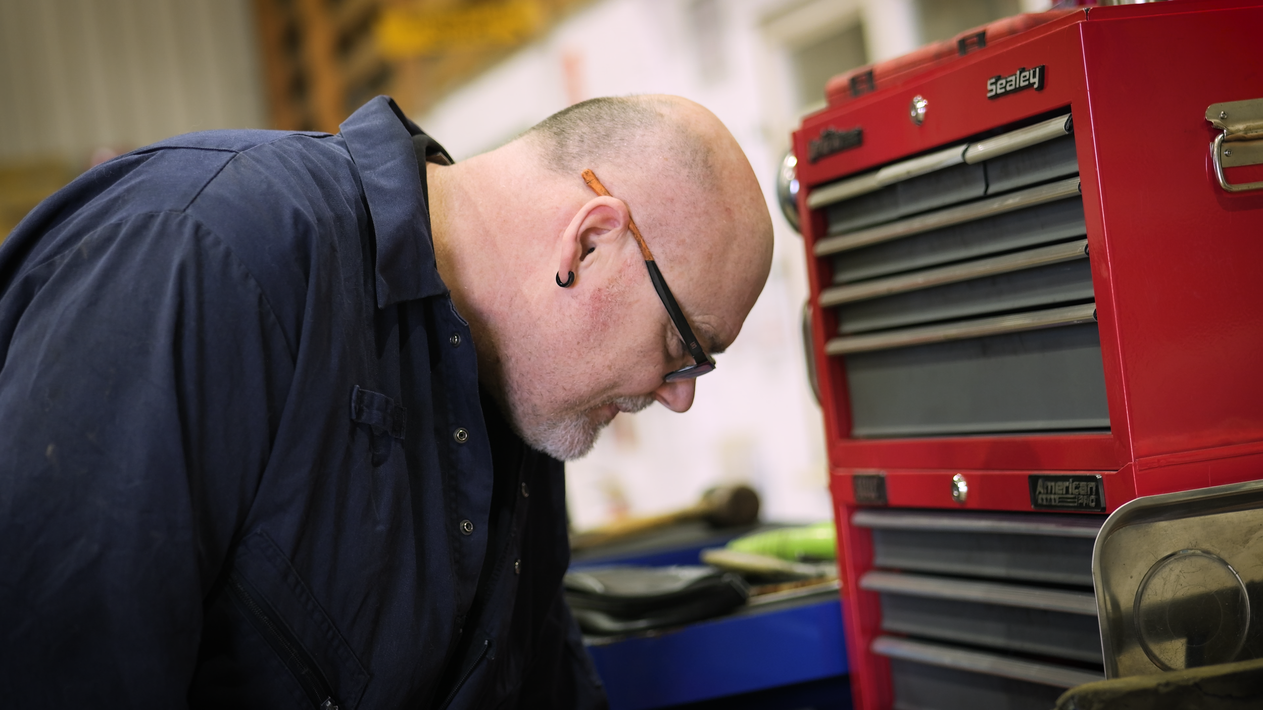 A man wearing glasses and a black jacket, leaning over a worktable, focused on a task, with a red tool chest nearby.