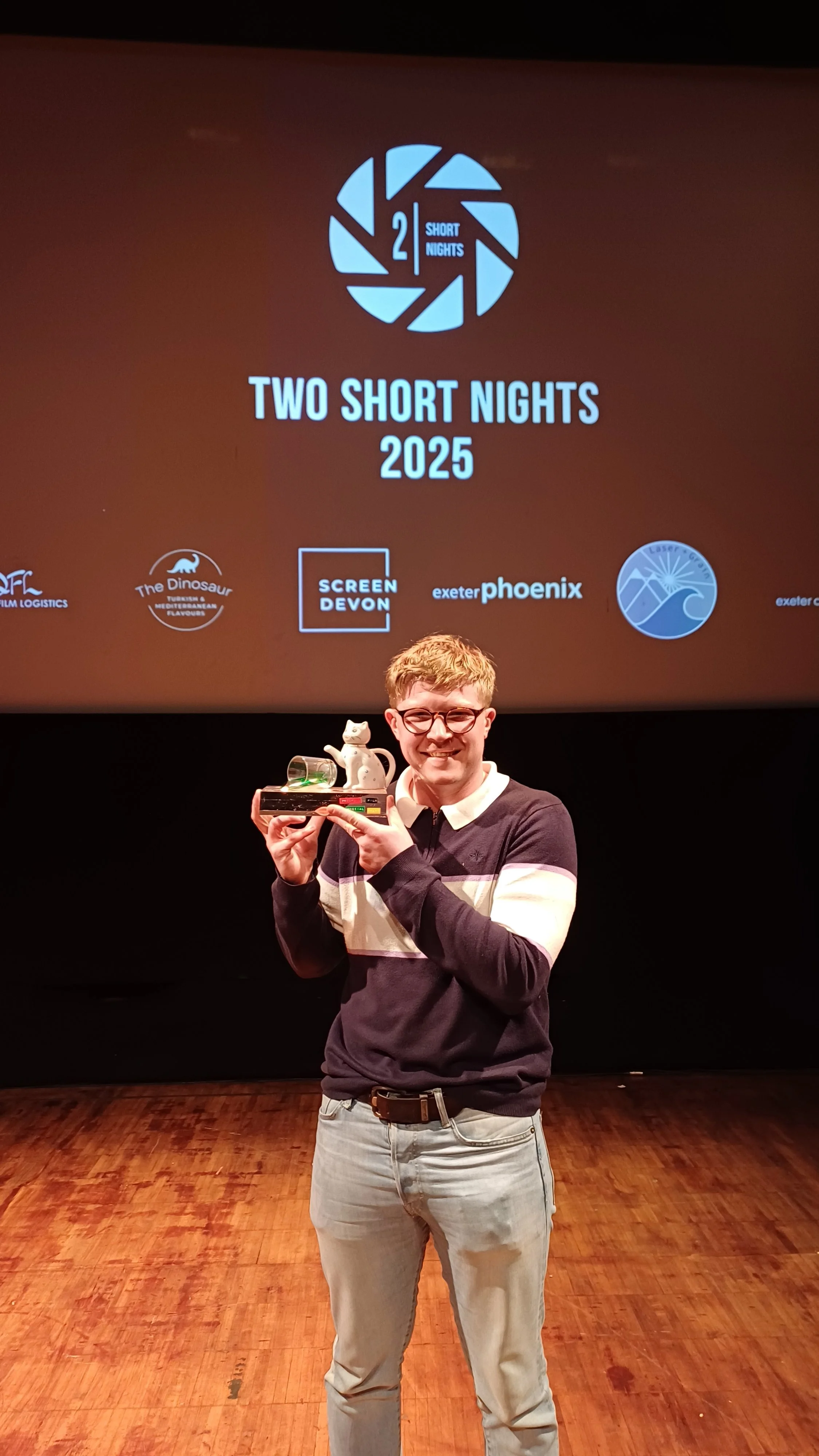 Young man with glasses, smiling, holding a trophy with a small cat figurine on top, standing in front of a projection screen displaying event details for 'Two Short Nights 2025' with logos of sponsors.