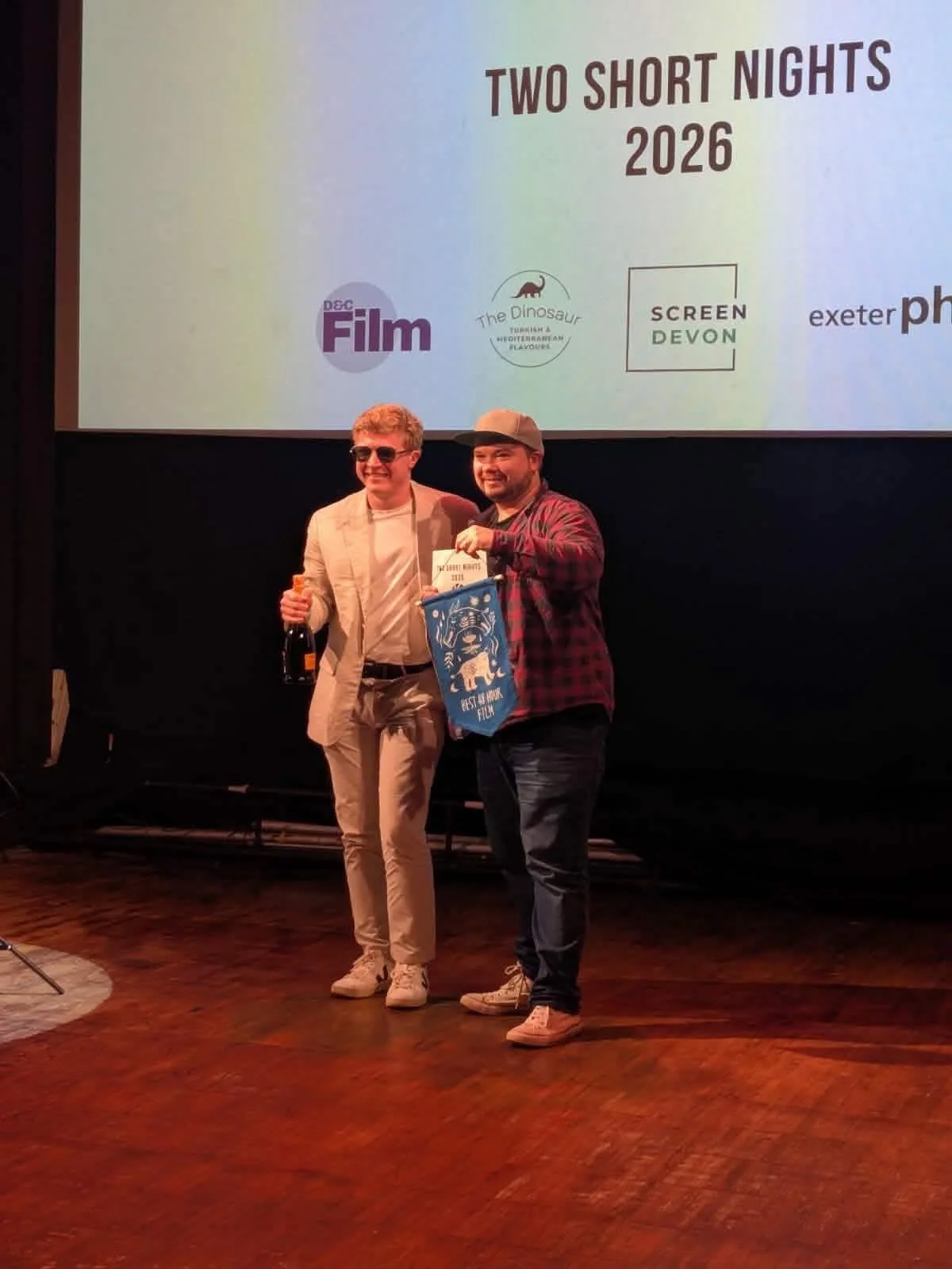 Connor Keal and Sam Cook smiling on stage at Two Short Nights awards ceremony, one holding a certificate and a blue flag, with a large screen behind them displaying event logos and the text 'Two Short Nights 2026'.