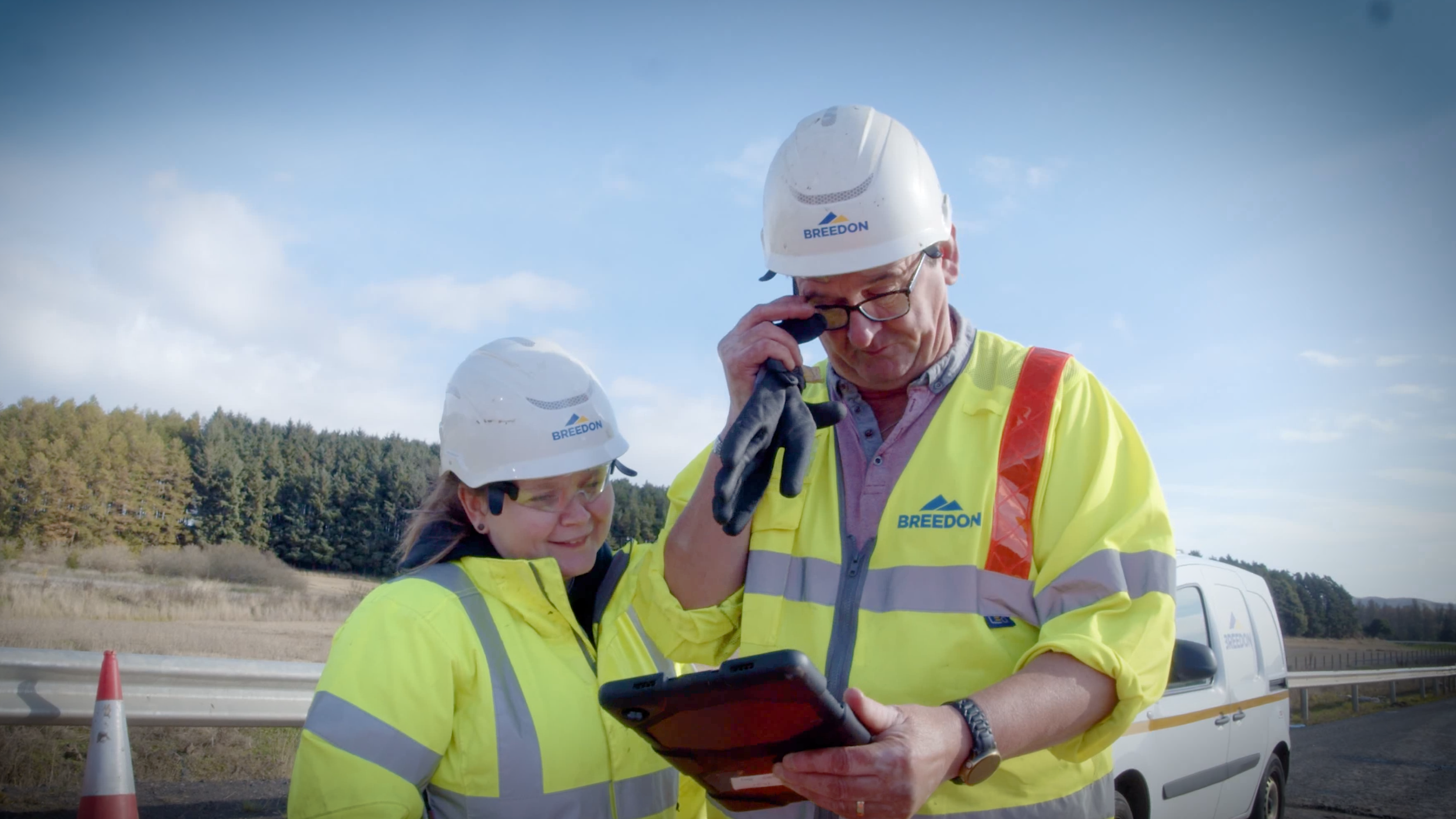 Two construction workers wearing yellow high-visibility jackets and white helmets labeled 'Breedon' standing outdoors next to a road, looking at a tablet, with a white van and a scenic landscape of trees and blue sky in the background.