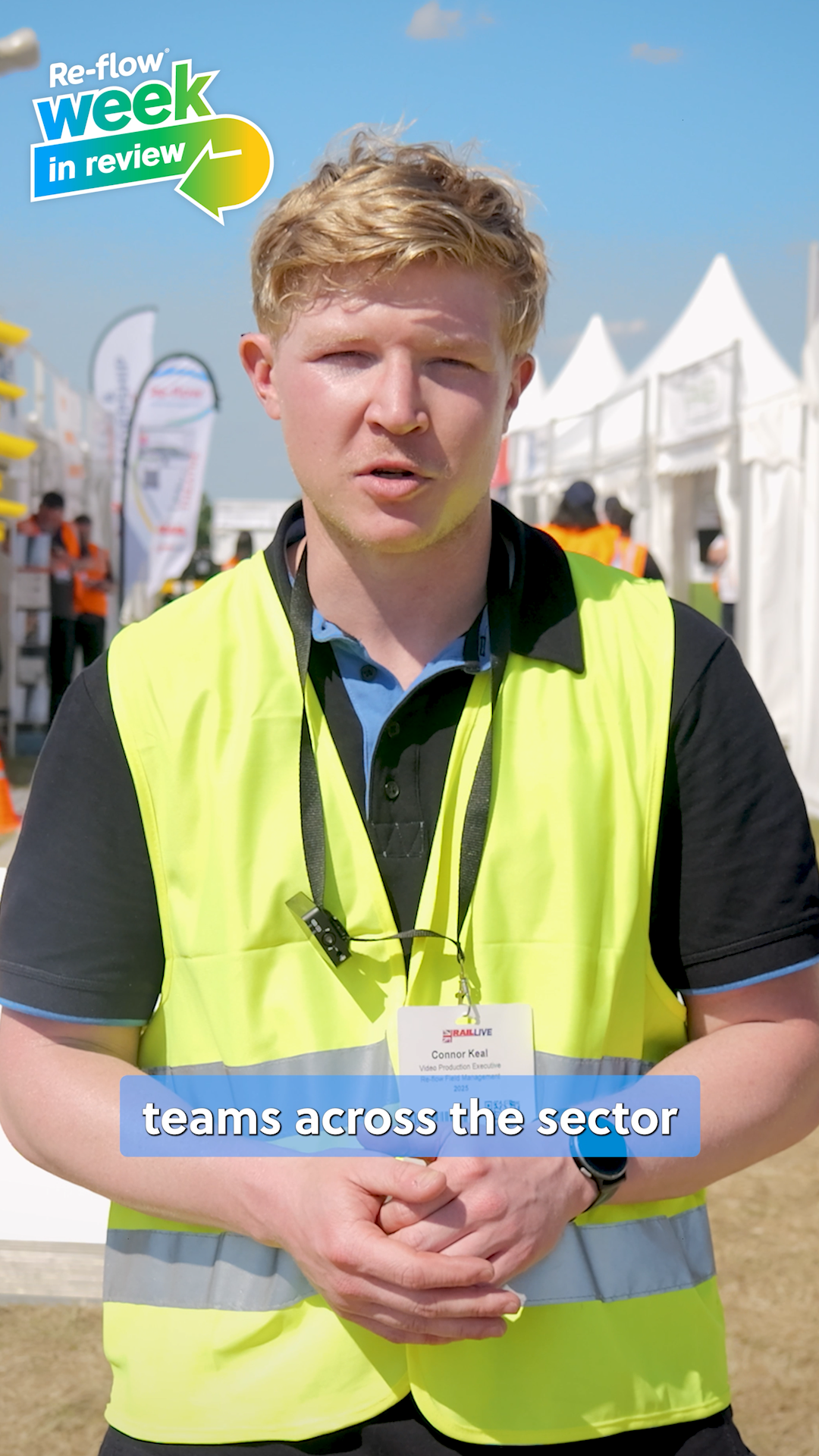 A man wearing a high-visibility yellow vest with a lanyard, standing outdoors at a race event with tents and banners in the background.