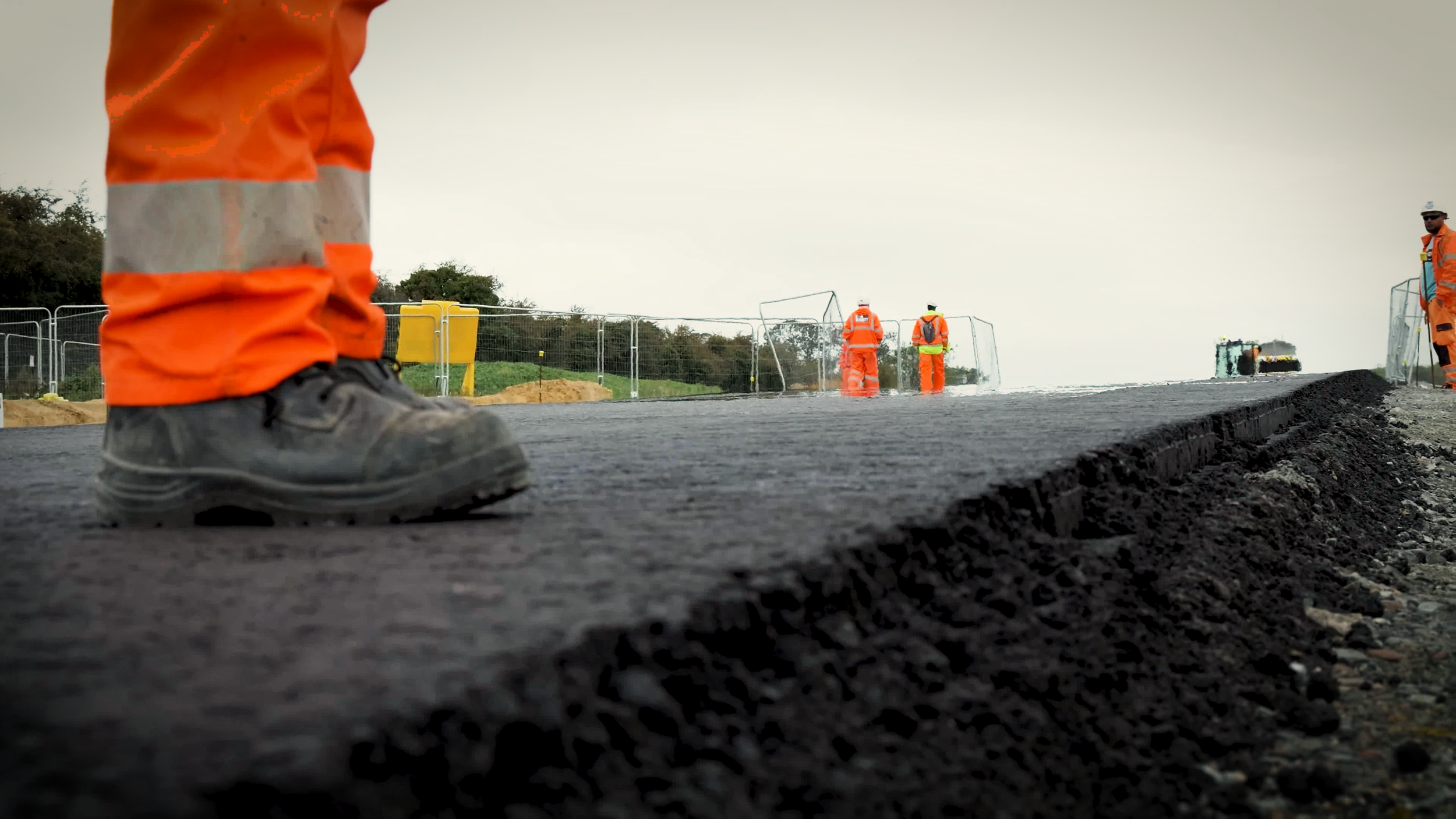 Close-up of a construction worker's boot standing on freshly paved asphalt with other workers in bright safety vestes and helmets working in the background.