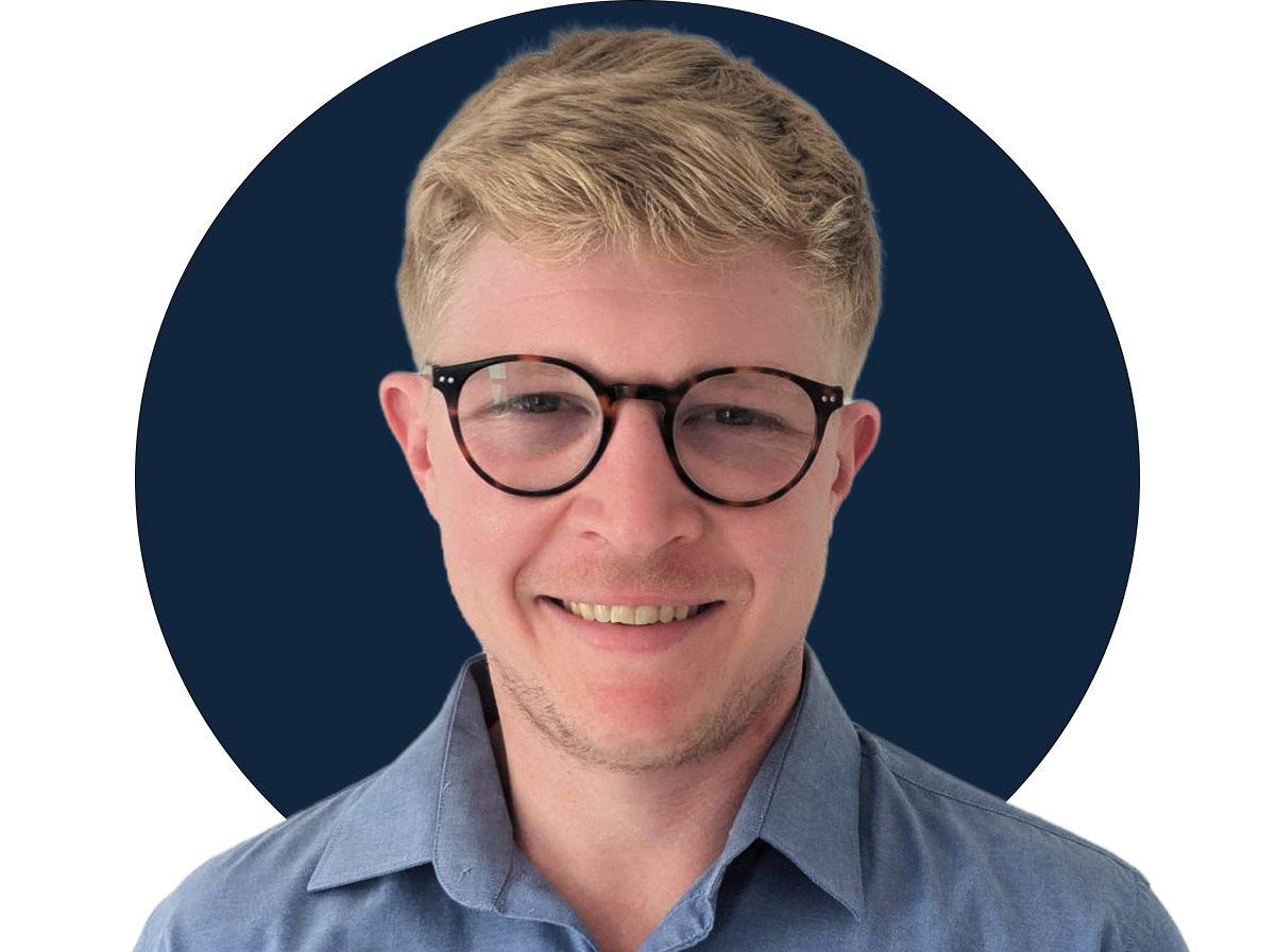 A young man with blonde hair, black glasses, and a light blue shirt smiling at the camera against a navy blue and black background.