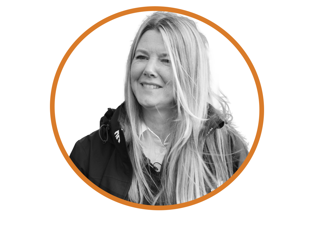 Black and white portrait of a smiling woman with long blonde hair, wearing a jacket and a collared shirt, set against an orange and black abstract background.
