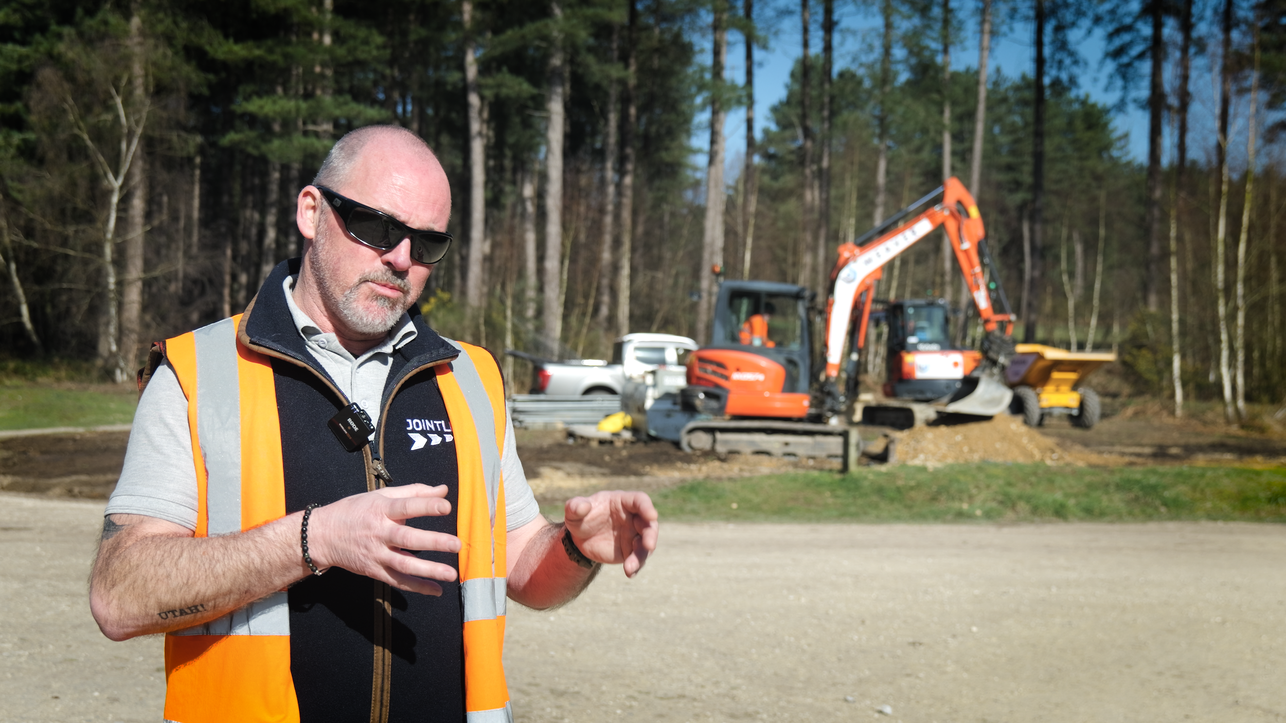 A construction worker wearing sunglasses and an orange safety vest stands outdoors in a wooded area with construction equipment, including a mini excavator and a dump trailer, in the background.