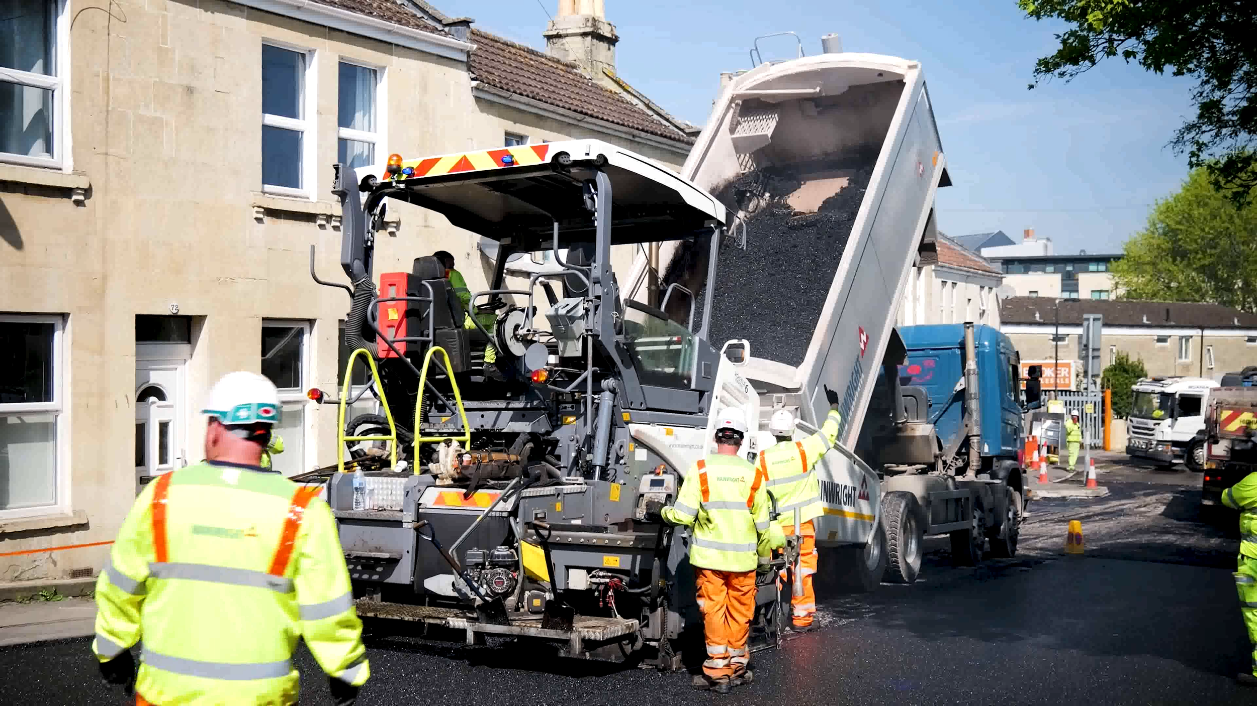 Construction workers in safety vests and helmets operating a large asphalt paving machine on a street, with a truck dumping asphalt into the machine, in a residential neighborhood.