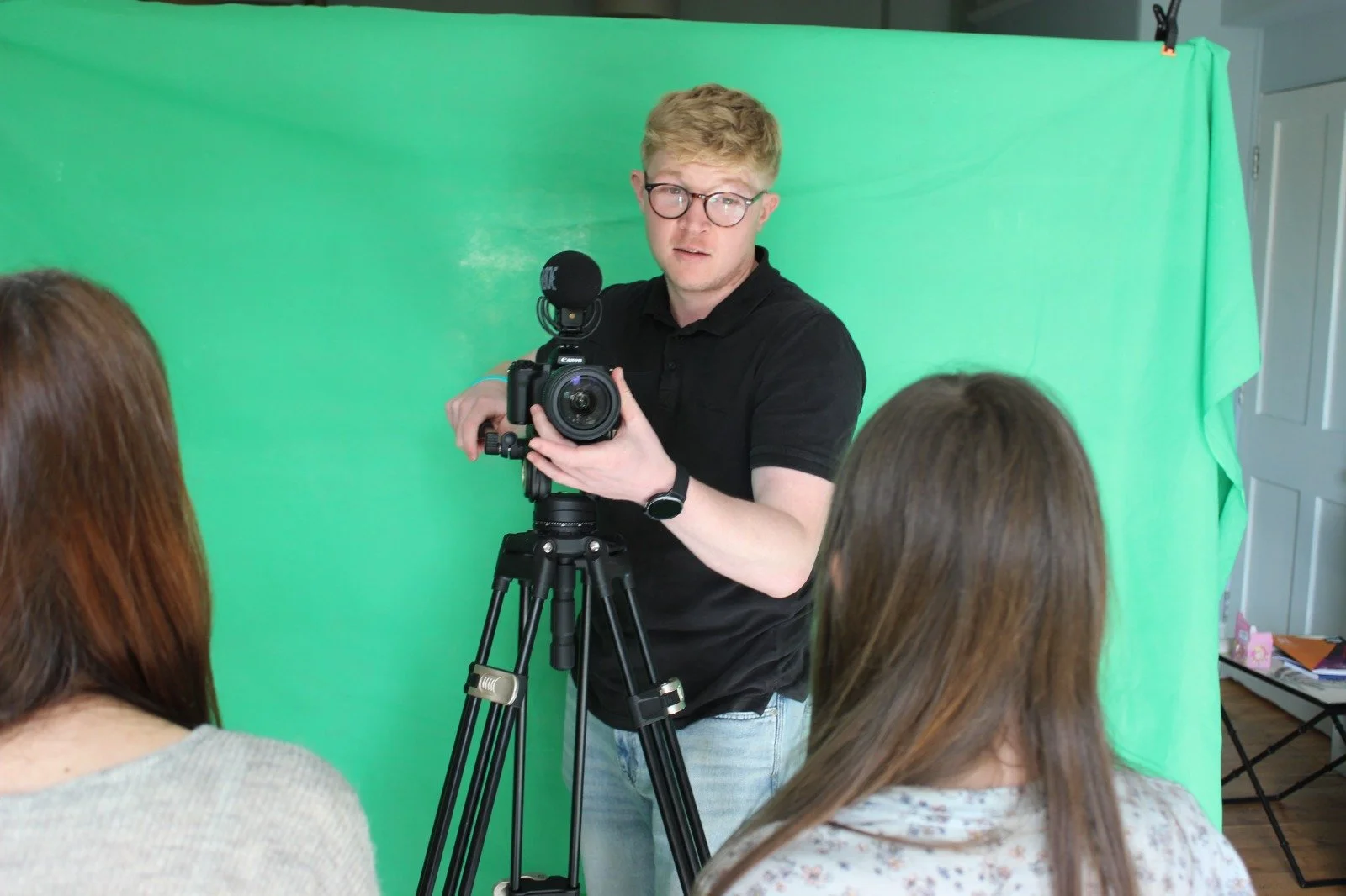 Connor Keal operating a professional camera on a tripod in front of a green screen, with two girls listening to him.