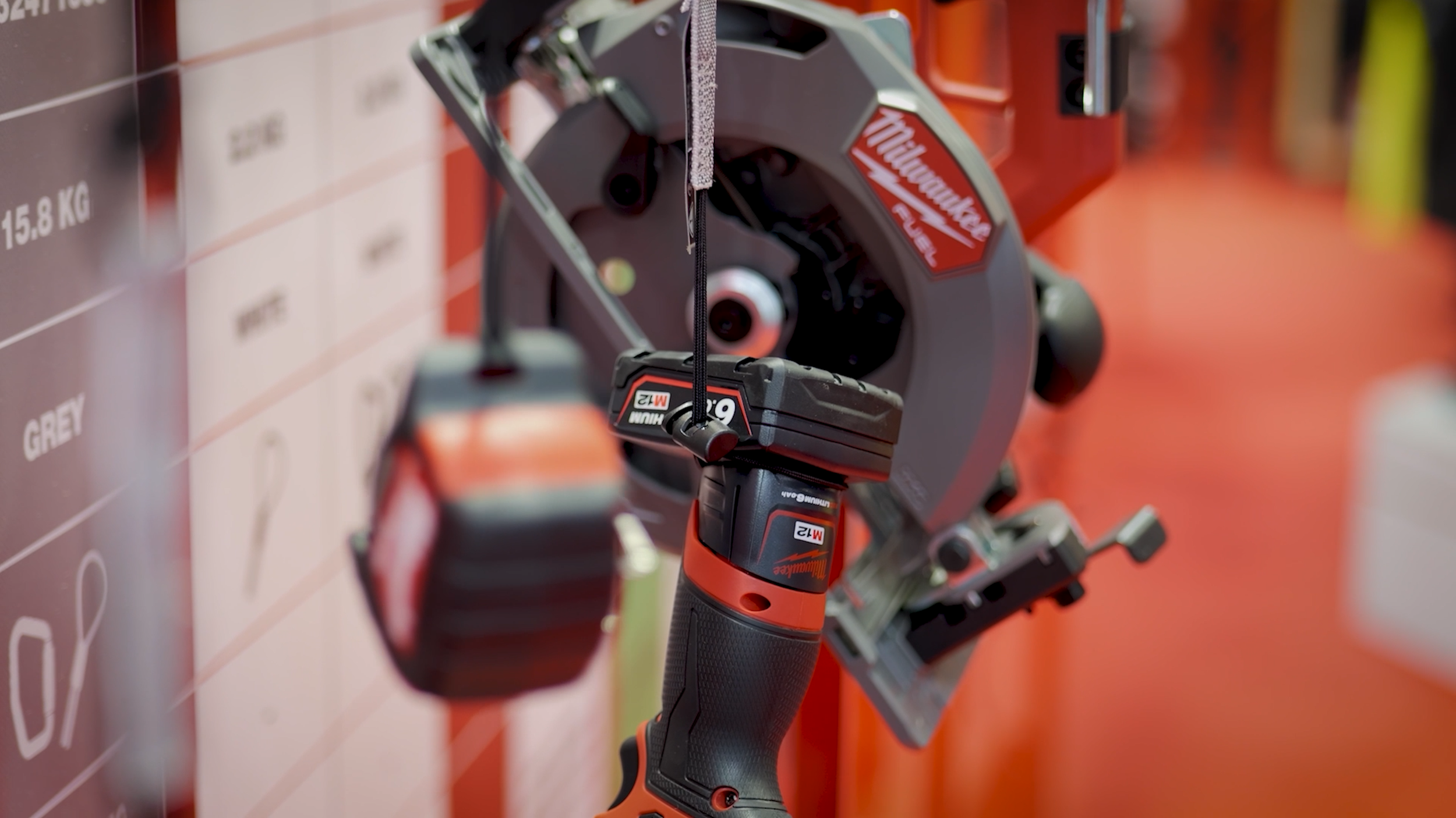 Close-up of a Milwaukee power tool with a chainsaw blade and a cordless drill with orange and black design, mounted on a display wall with blurred item labels.