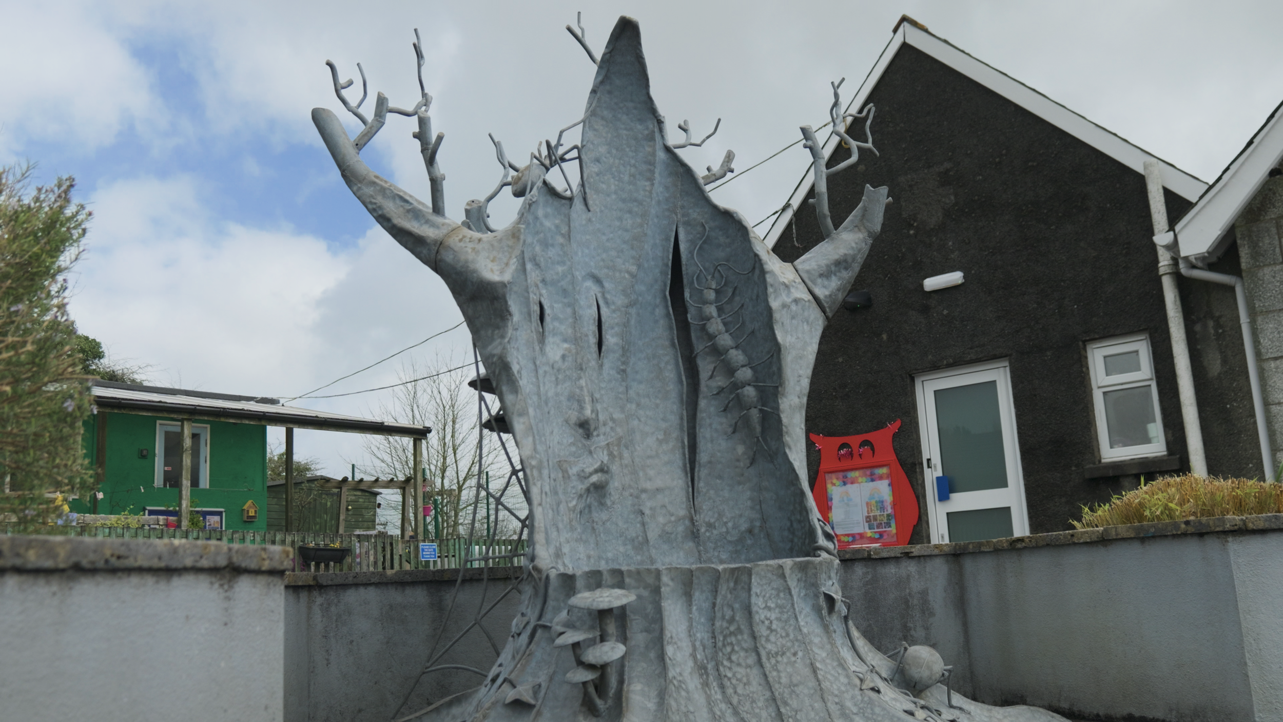 A metallic sculpture of a fairy tale character with a pointed hat and mystical features is situated in a garden area, near a black house with white windows and a children's playhouse.