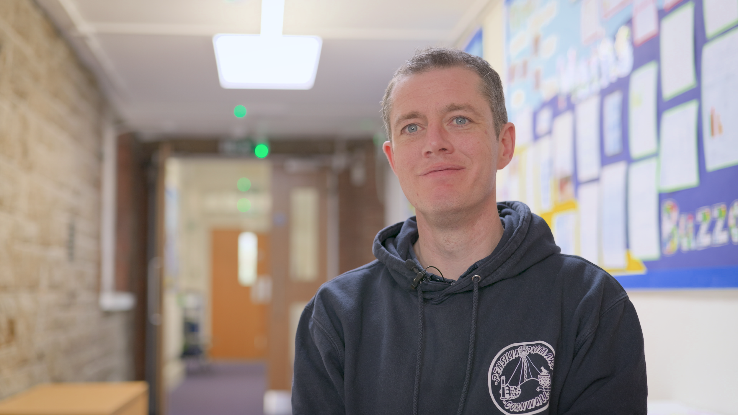 A man with short gray hair, blue eyes, and wearing a black hoodie with a logo, standing in a hallway with bulletin boards on the wall behind him.