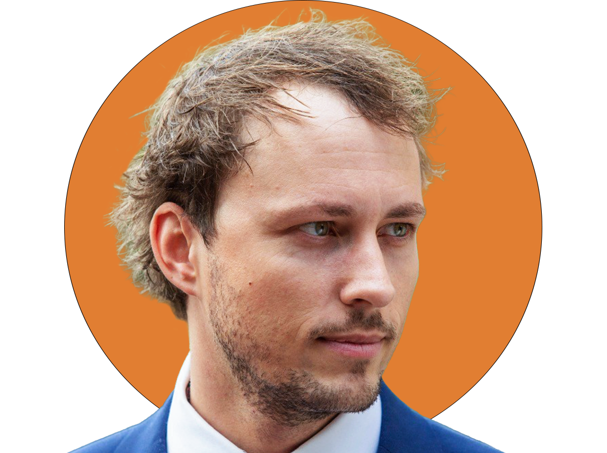 Close-up of a young man with light brown hair, slight beard, wearing a formal suit, against an orange background.