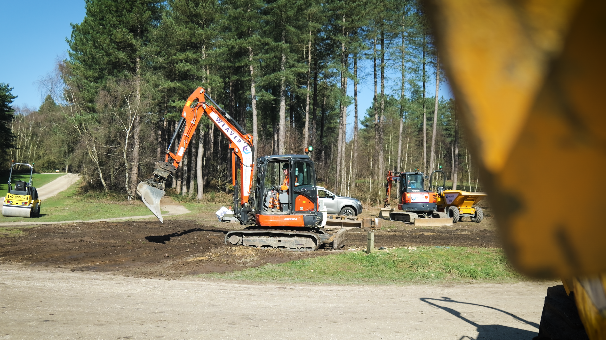 A construction site with a small orange excavator operated by a person in orange safety gear, a yellow roller compactor, and a small yellow dump truck, with trees and a clear blue sky in the background.