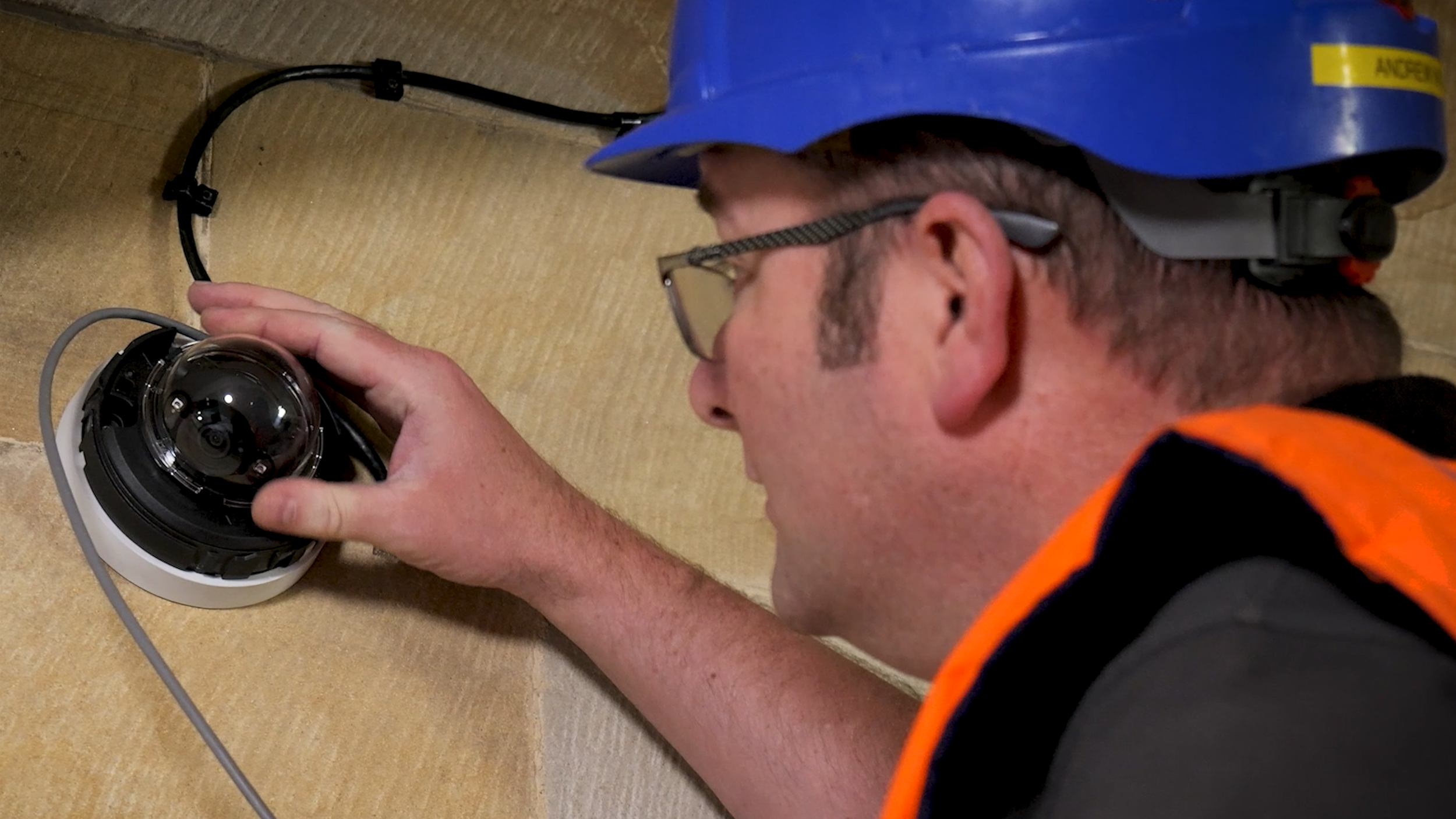 A worker wearing a blue safety helmet and glasses, dressed in a black shirt with orange safety vests, installing or inspecting a security camera on a beige wall.