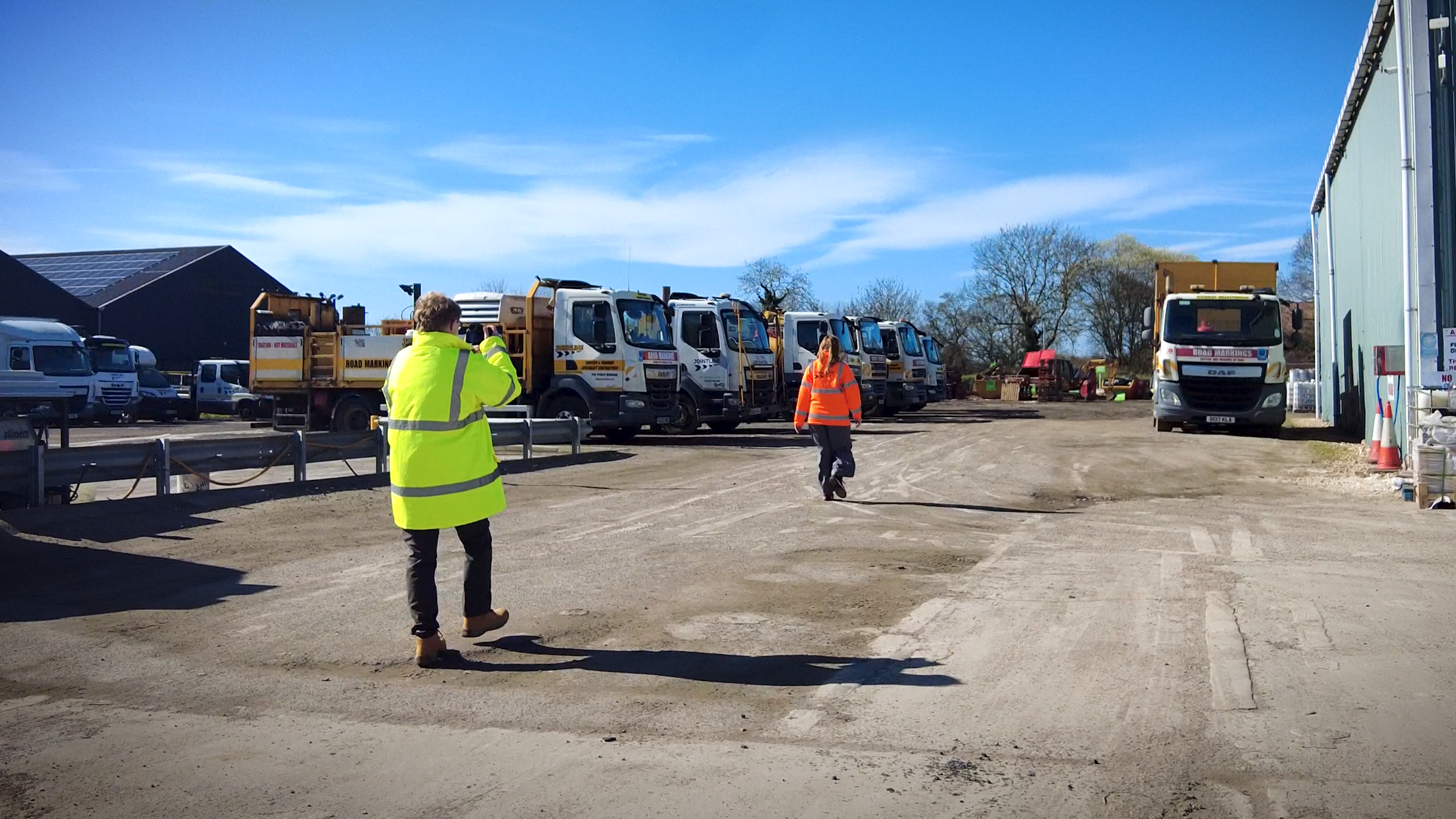 People in safety vests walking on a dirt lot with multiple trucks parked nearby, under a clear blue sky.