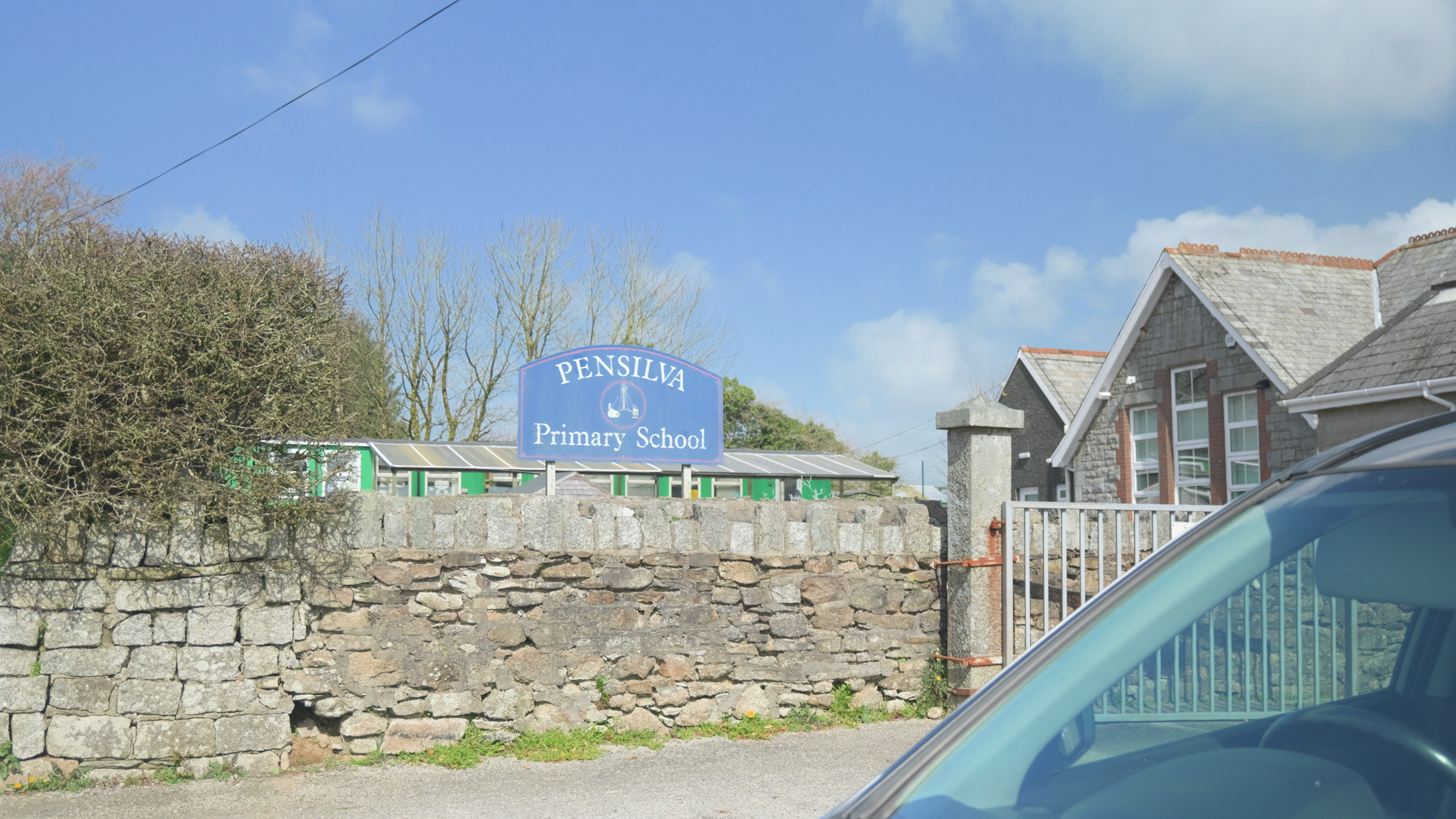 Sign for Pensilva Primary School with stone wall, houses, and trees in the background under a partly cloudy sky.