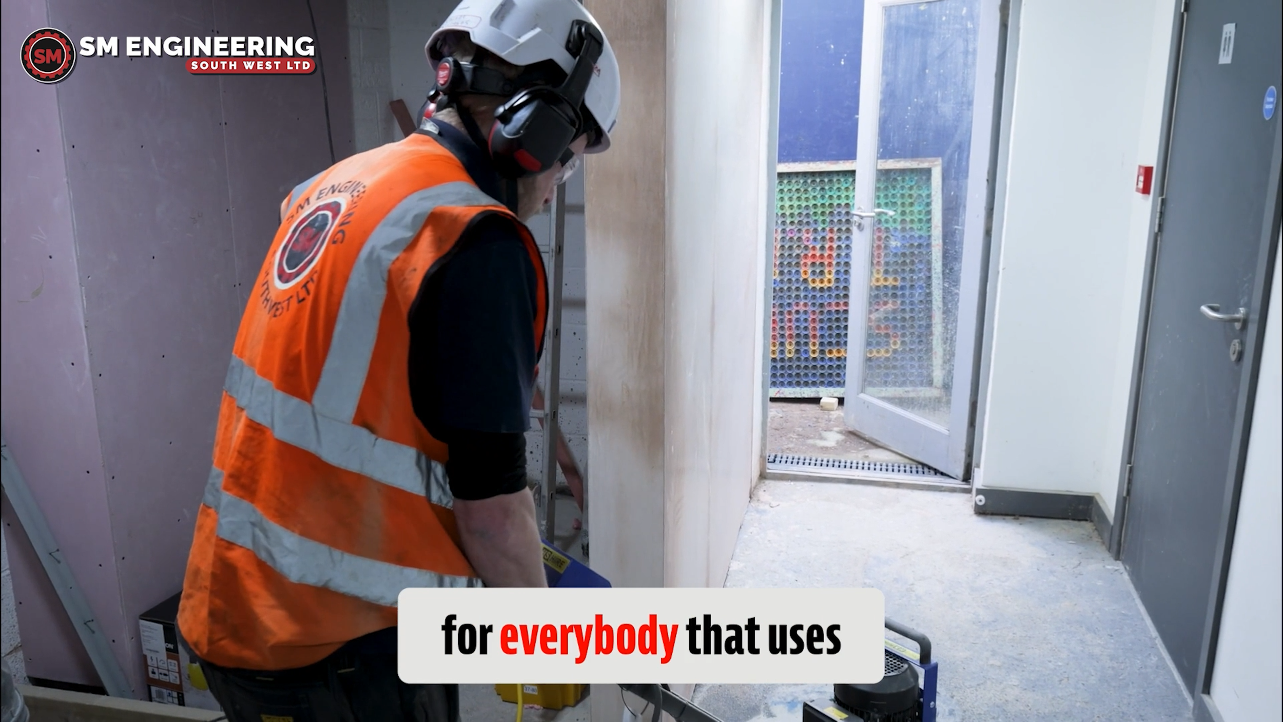 A worker wearing an orange safety vest and a white helmet with a headset inside an industrial or construction area. There is a door on the right side and machinery on the floor.