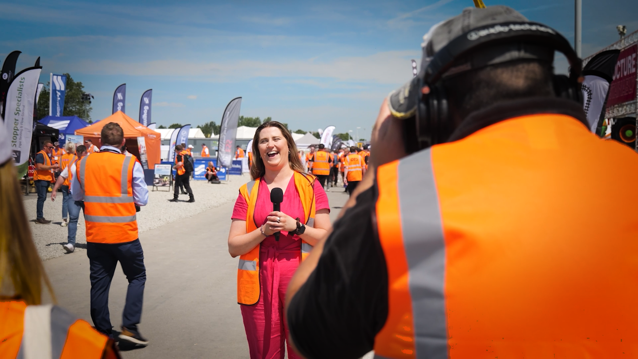 A woman in a pink dress and orange safety vest is smiling and speaking into a microphone while being interviewed at an outdoor event. People in orange safety vests are walking and standing around, with tents and flags in the background on a sunny day.