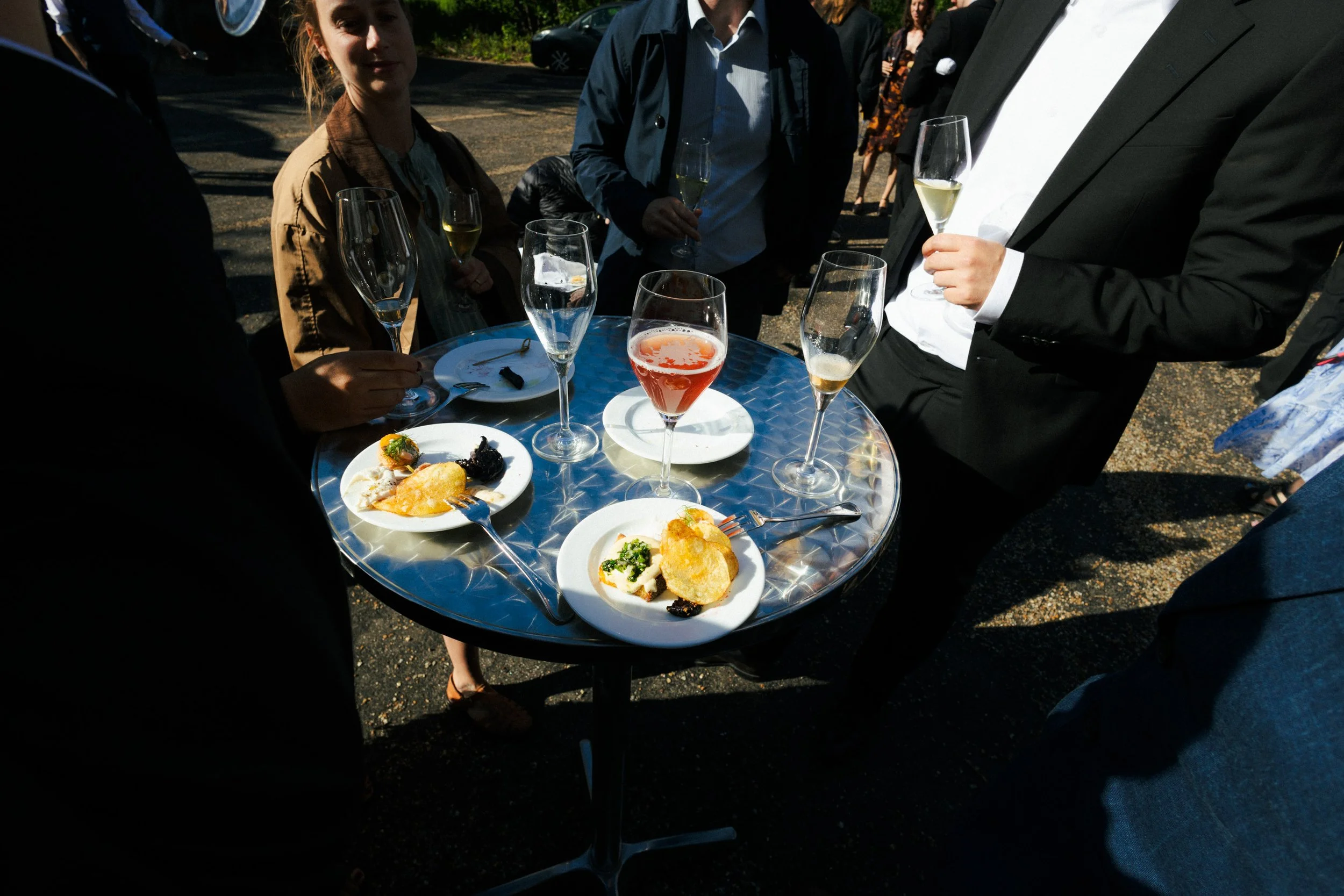 En gruppe mennesker ved en udendørs middag med tallerkener med mad og glas med vin eller champagne, på en rund metalbord. Mennesker i formelle og afslappede tøj, sollys og skygge. Baggrund med grønne buske og en asfaltvej.