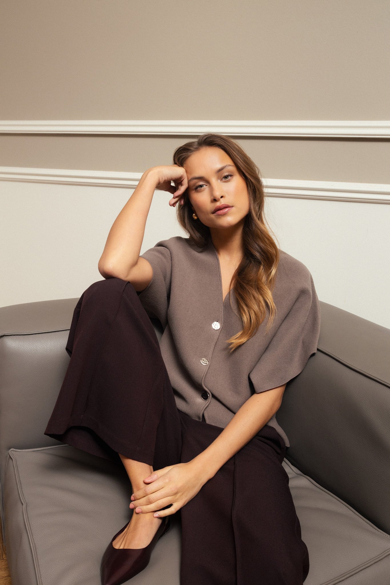A woman with long wavy hair sitting on a gray sofa, resting her head on her hand, wearing a taupe button-up shirt and black pants, with her legs crossed and wearing black heels, in a room with a beige wall and white trim.