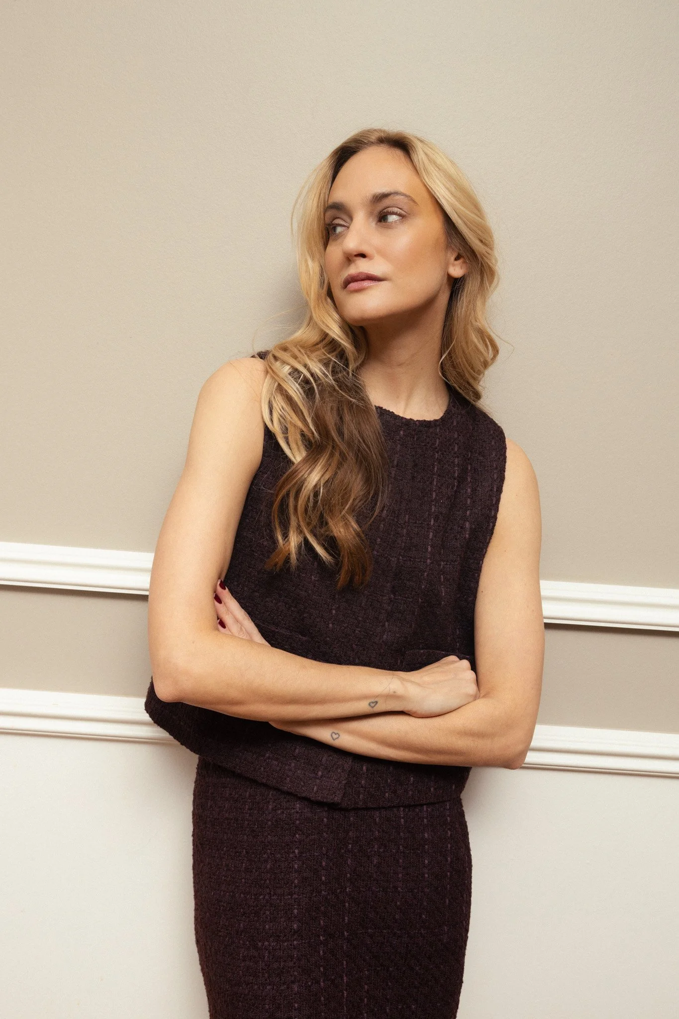 A woman with long wavy hair, wearing a sleeveless dark dress, standing against a plain beige wall with white molding, looking to her left with arms crossed.