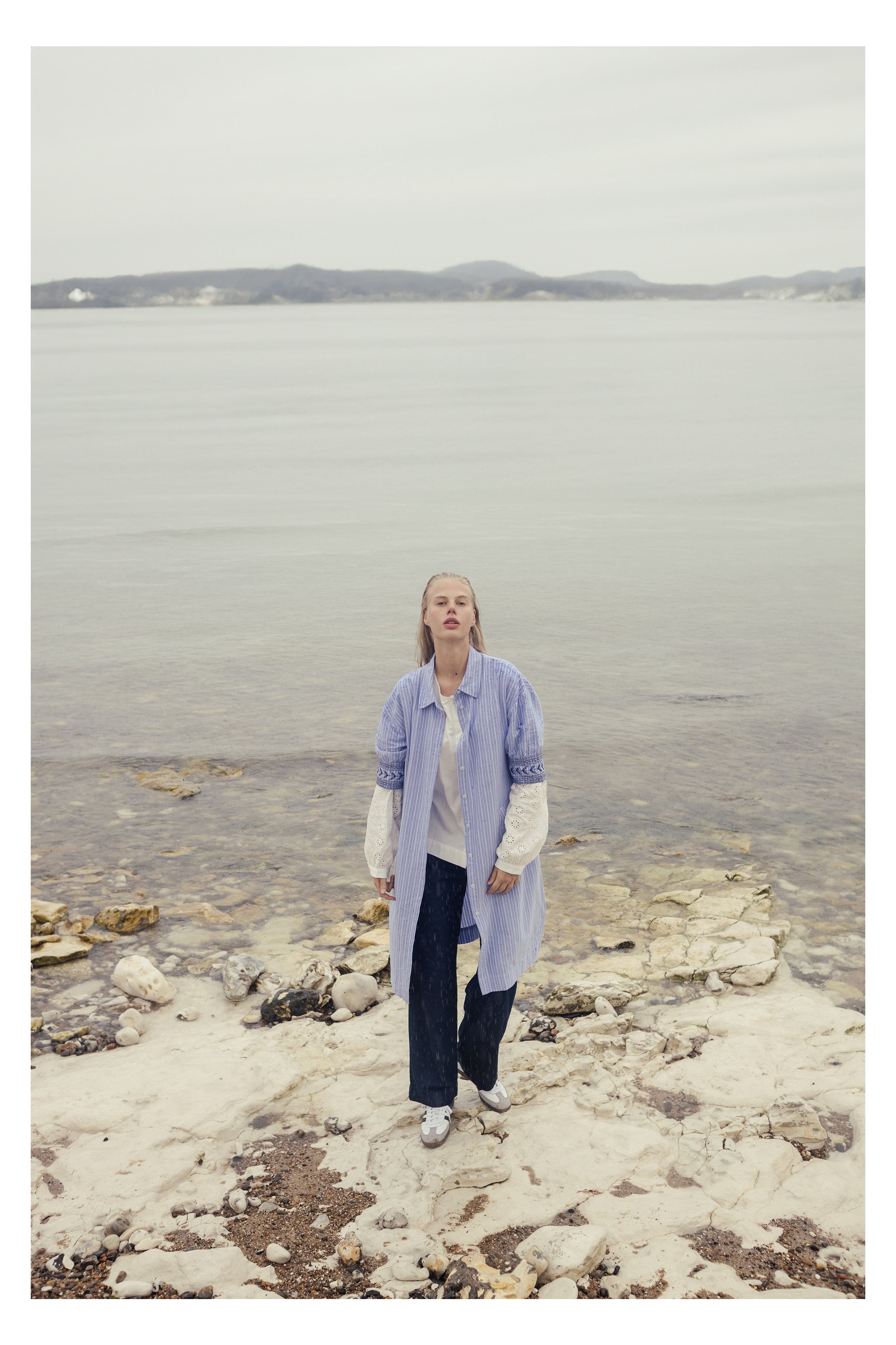 A young woman with blonde hair standing on a rocky and sandy shoreline, wearing a blue and white striped long shirt, black pants, and white sneakers, with a calm body of water and distant hills in the background.