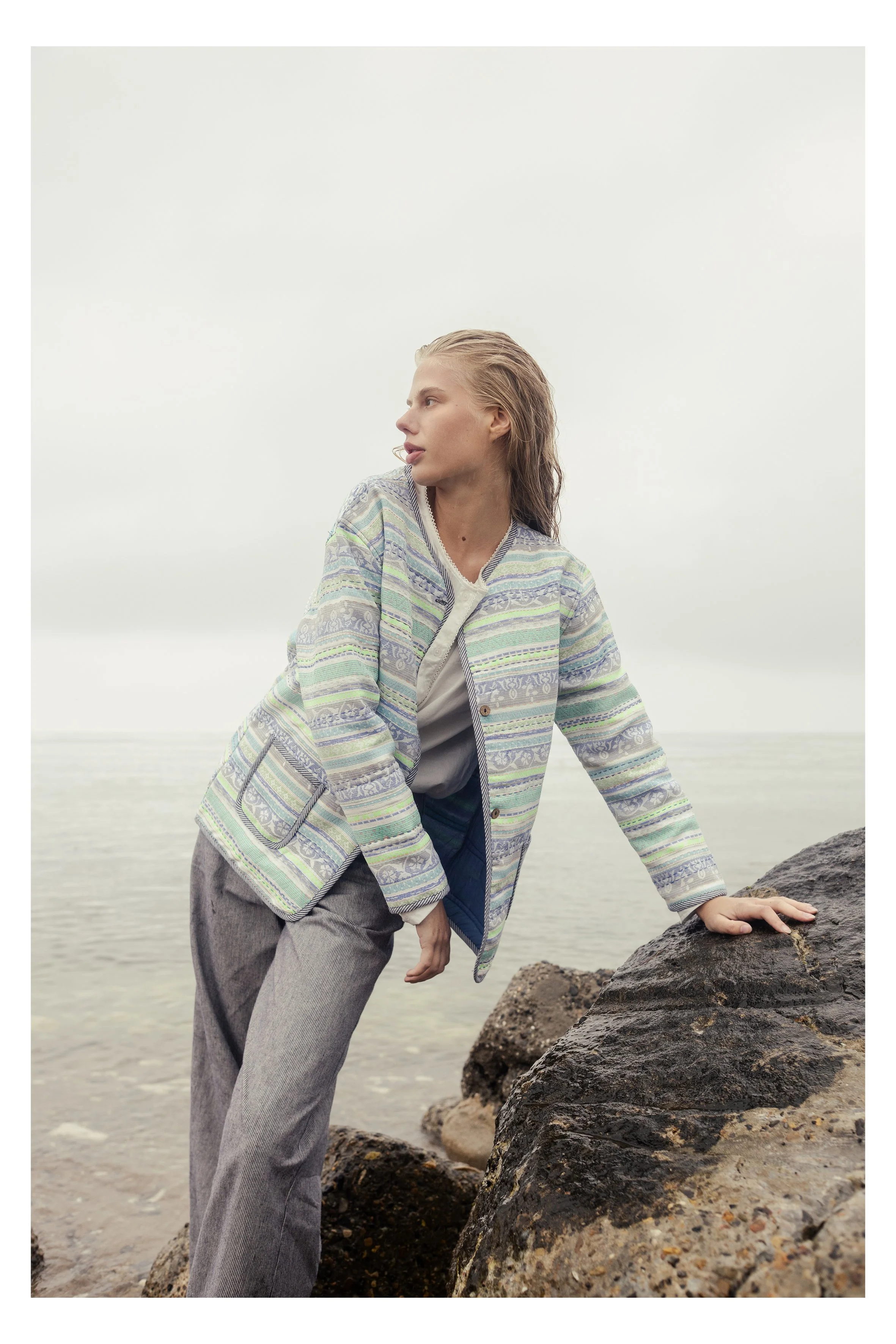 A woman with wet hair wearing a patterned jacket and gray pants, leaning on rocks by the water, with an overcast sky and calm ocean in the background.