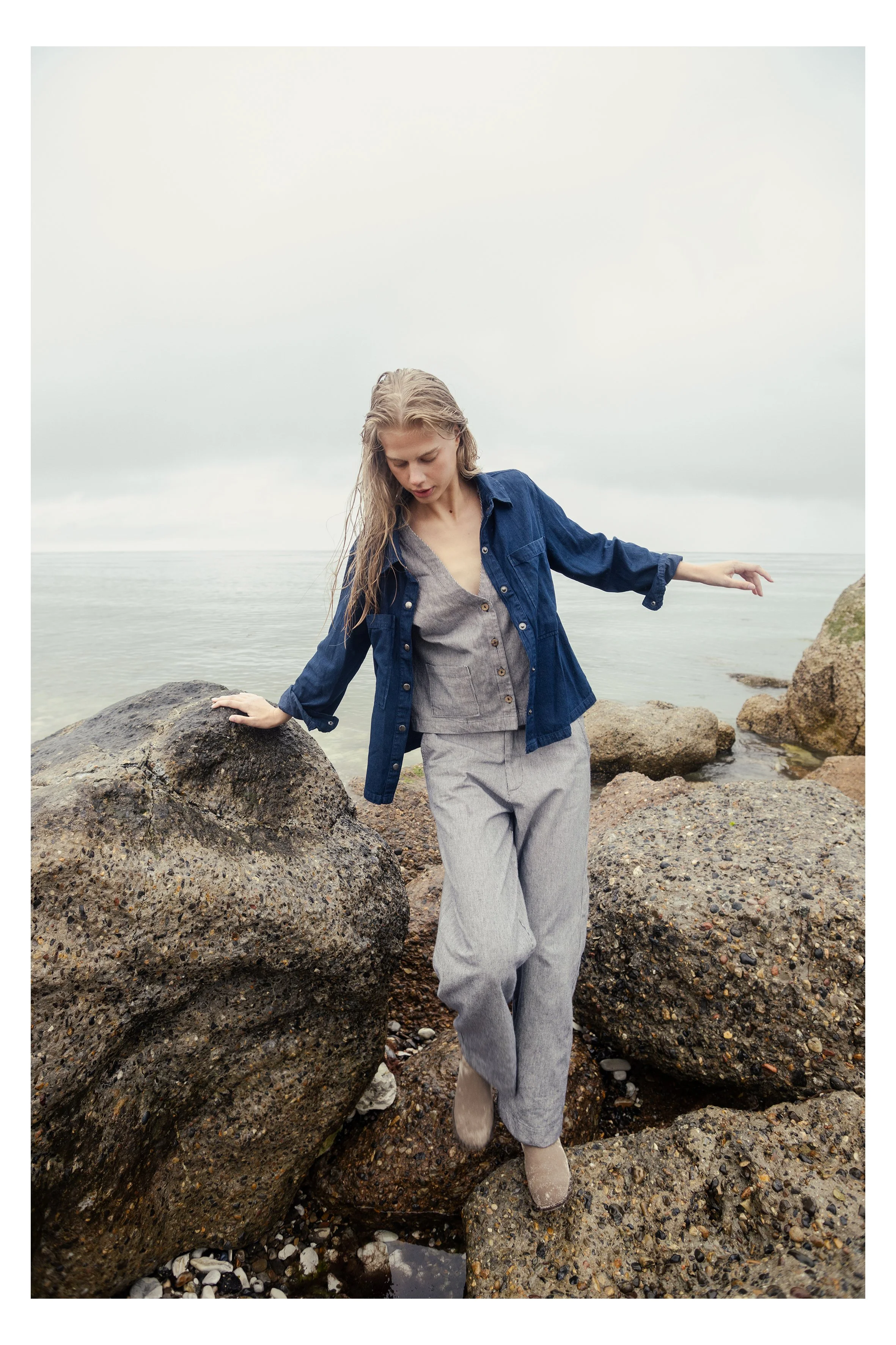A woman in a denim jacket and gray striped pants standing on rocks by the shoreline with water and overcast sky in the background.