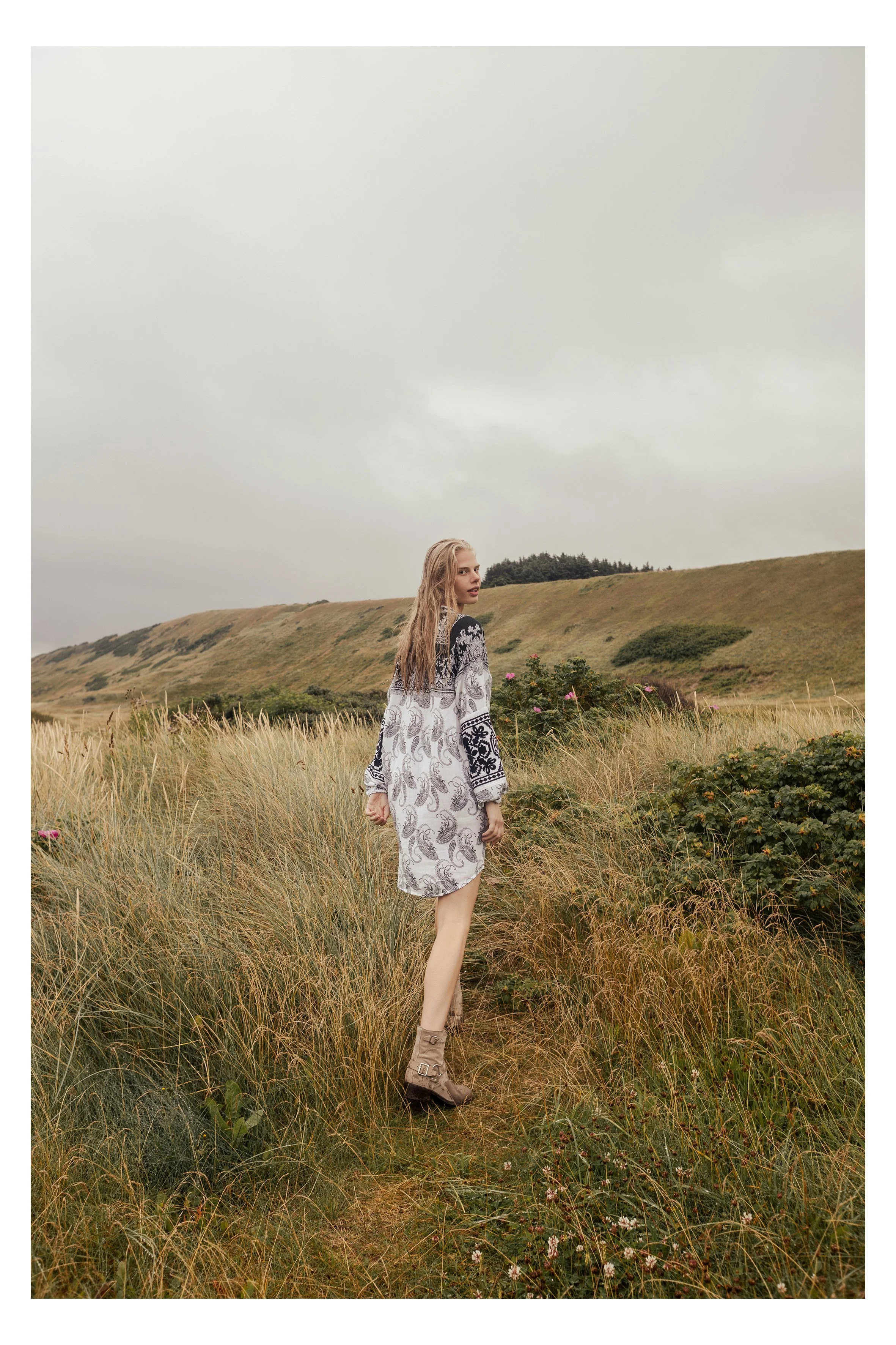 A young woman with long blonde hair wearing a white dress with black patterns and beige ankle boots walking through a grassy field with a hill and cloudy sky in the background.