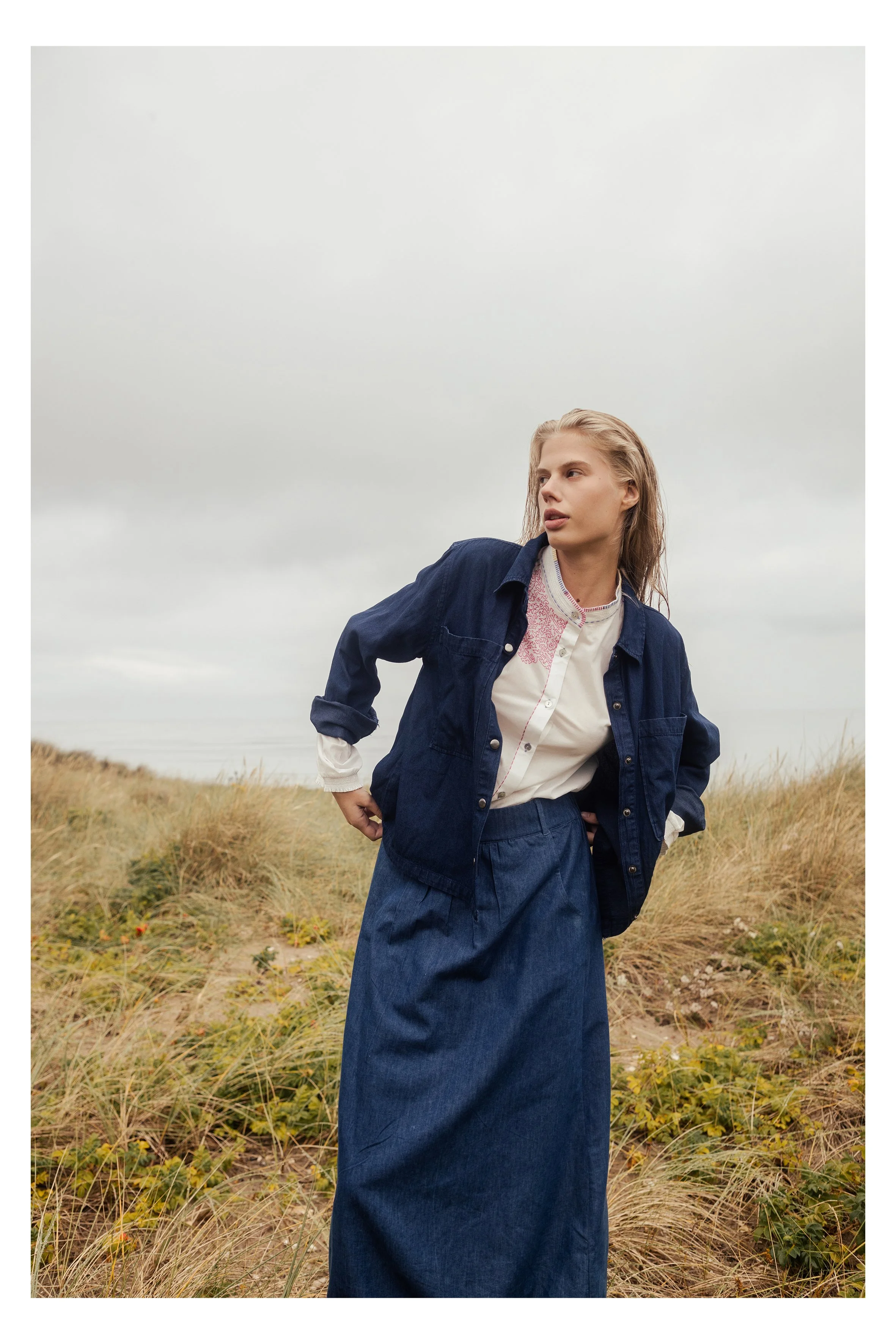 A woman with light hair standing in a grassy field under a cloudy sky, dressed in a white shirt and a blue denim jacket, with her hands on her hips, looking to the side.