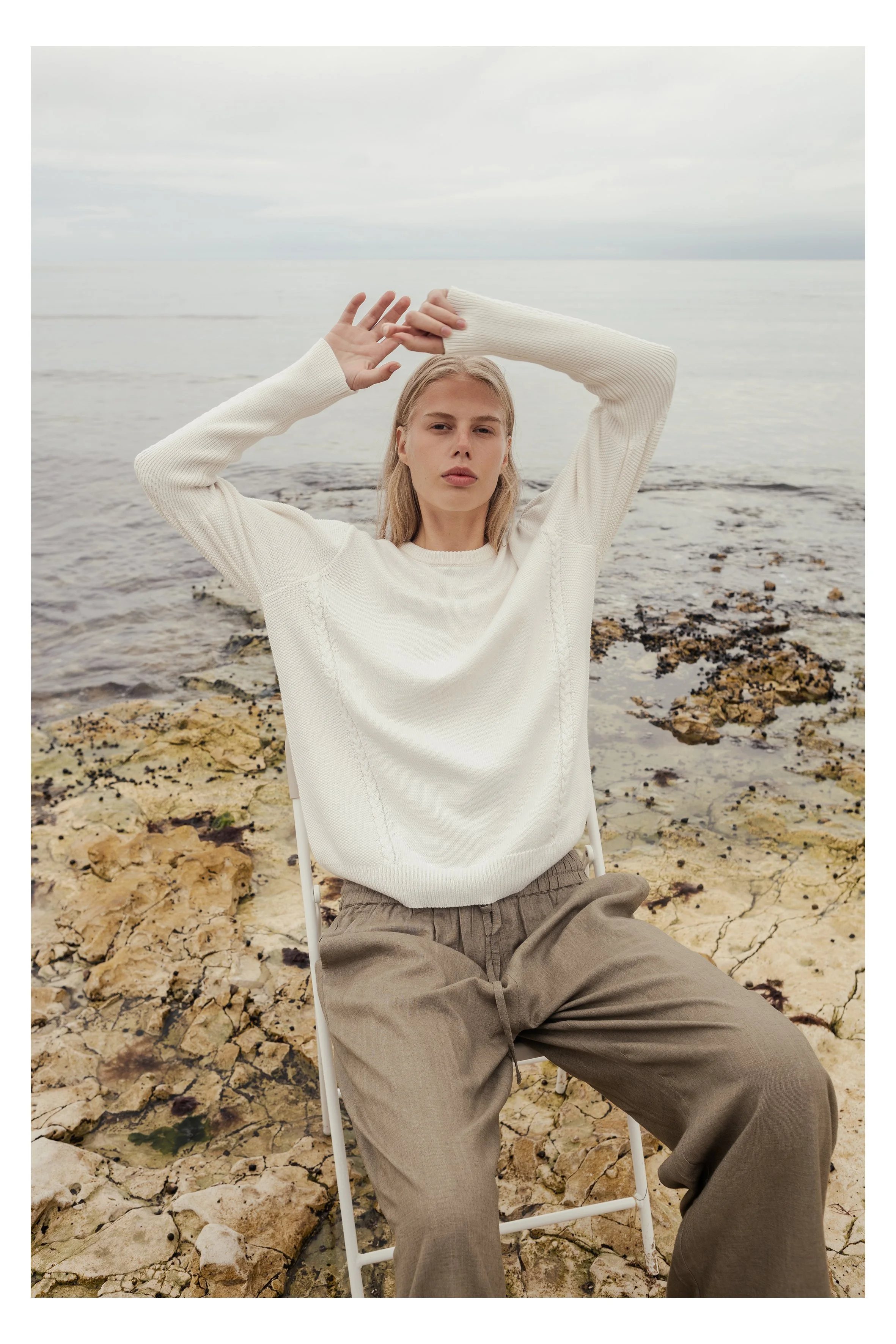 A young woman with blonde hair and neutral expression sitting on a white chair near the water on a rocky beach, wearing a cream sweater and loose brown pants, with her arms raised above her head.
