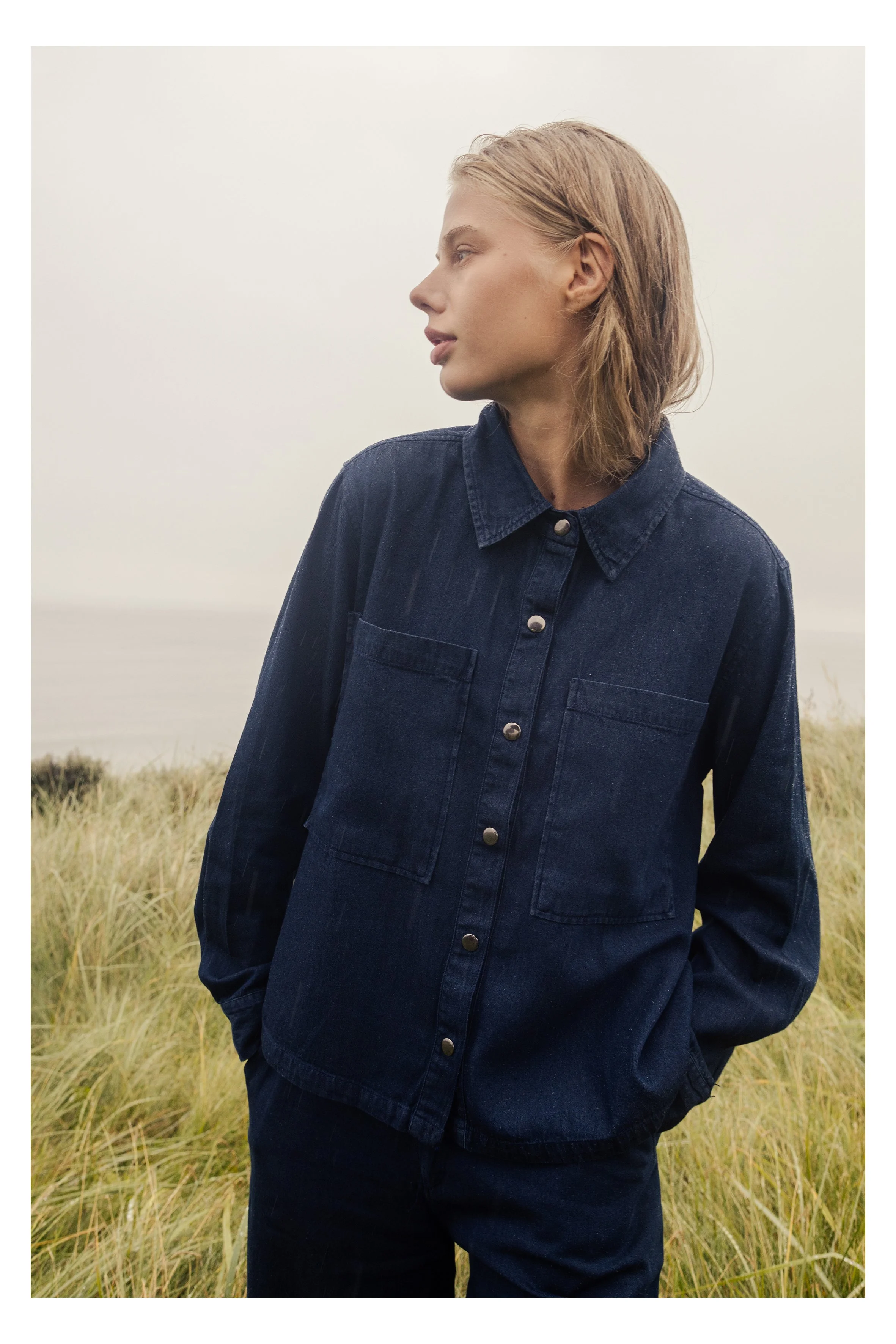 A woman with blond, wavy hair is standing outdoors in a field of tall grass, wearing a dark blue denim jacket and pants, looking to the left with a neutral expression.