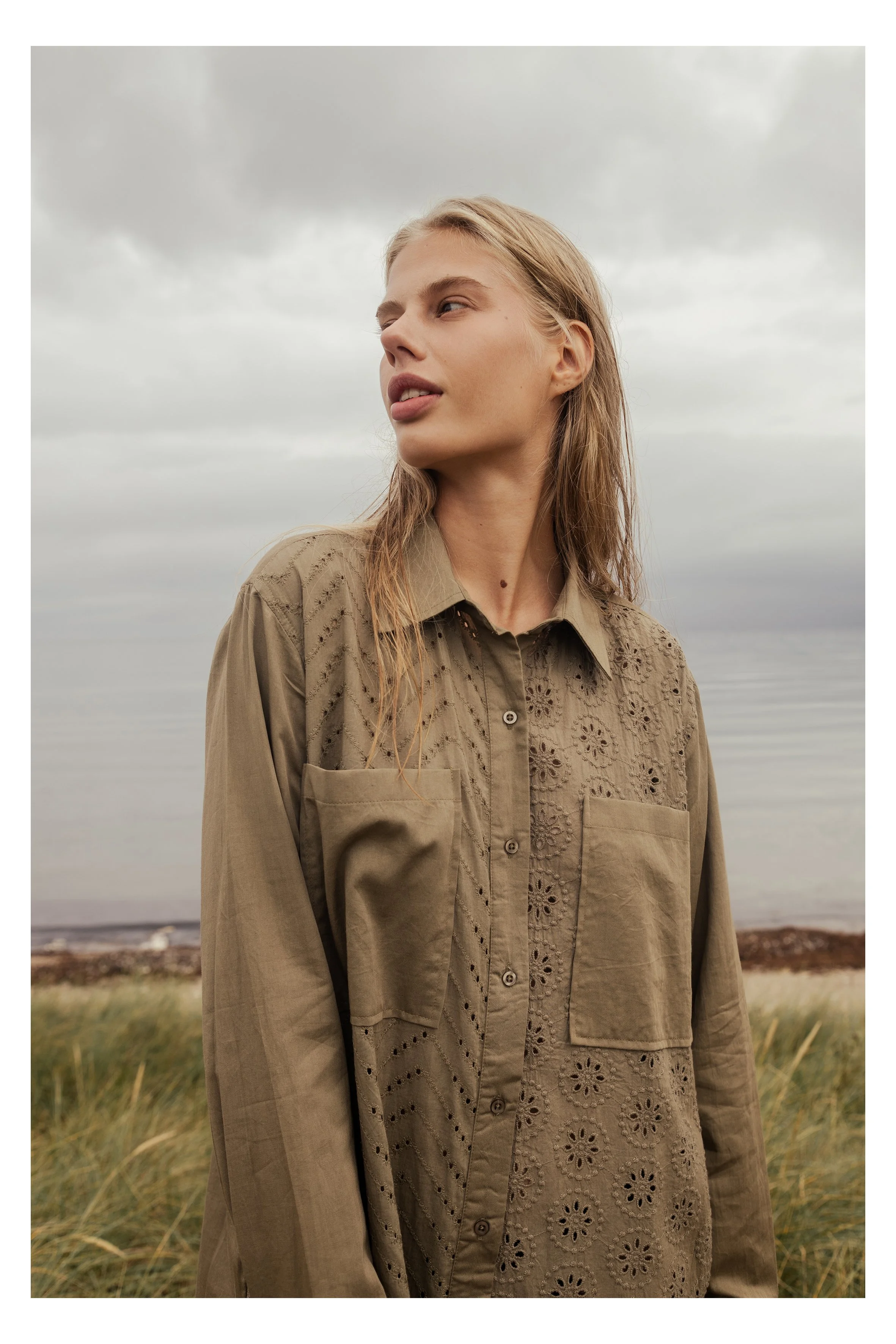 A woman with long blonde hair, wearing a tan button-up shirt with eyelet embroidery, is standing outdoors on a cloudy day near a body of water, with grass and a shoreline in the background.