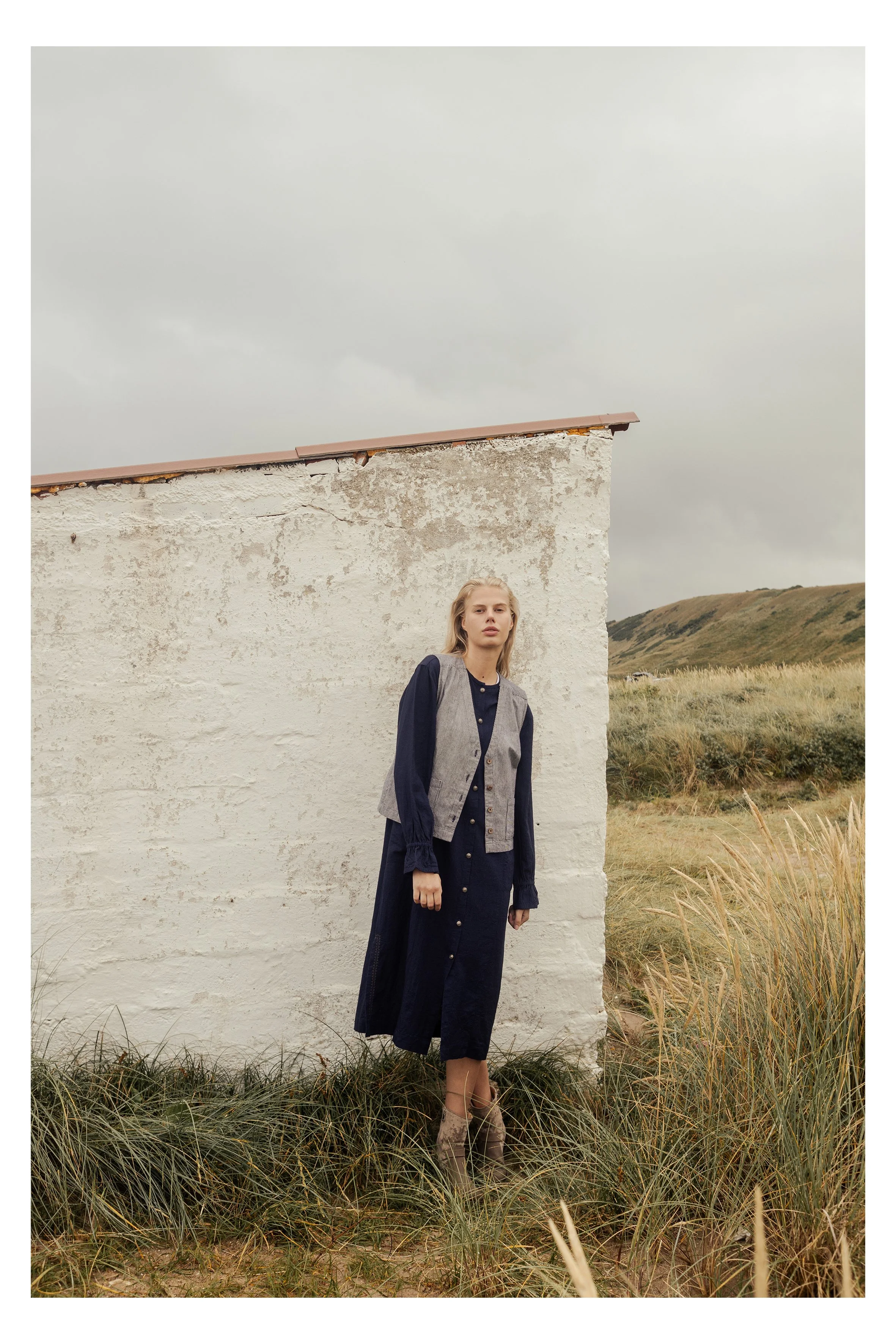 A woman standing outdoors in front of a weathered, white stucco building with a grassy field and rolling hills in the background, wearing a dark dress and a gray vest, with tall boots.