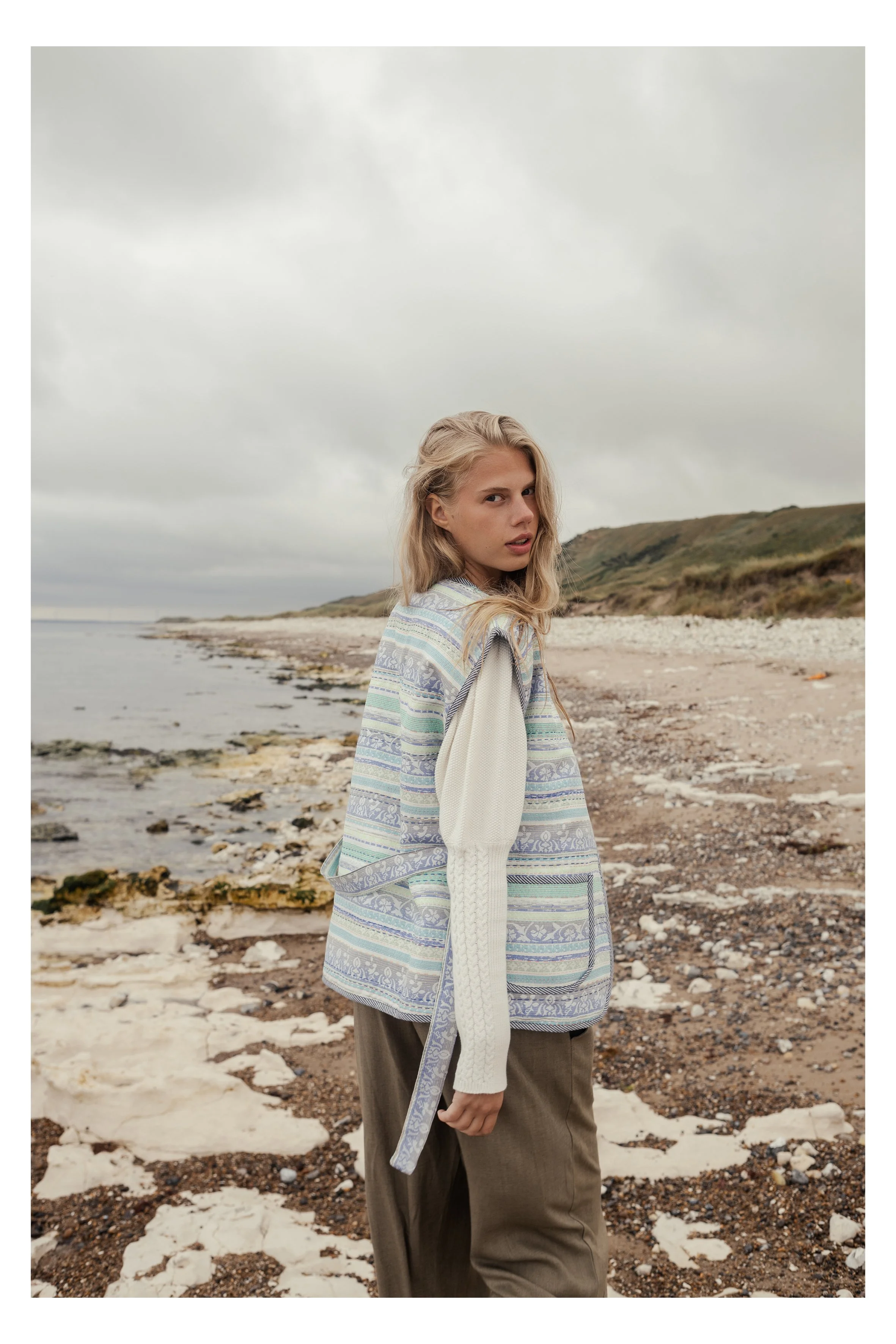 A woman with blonde hair standing on a rocky beach with ocean and hills in the background, wearing a patterned coat and brown pants.