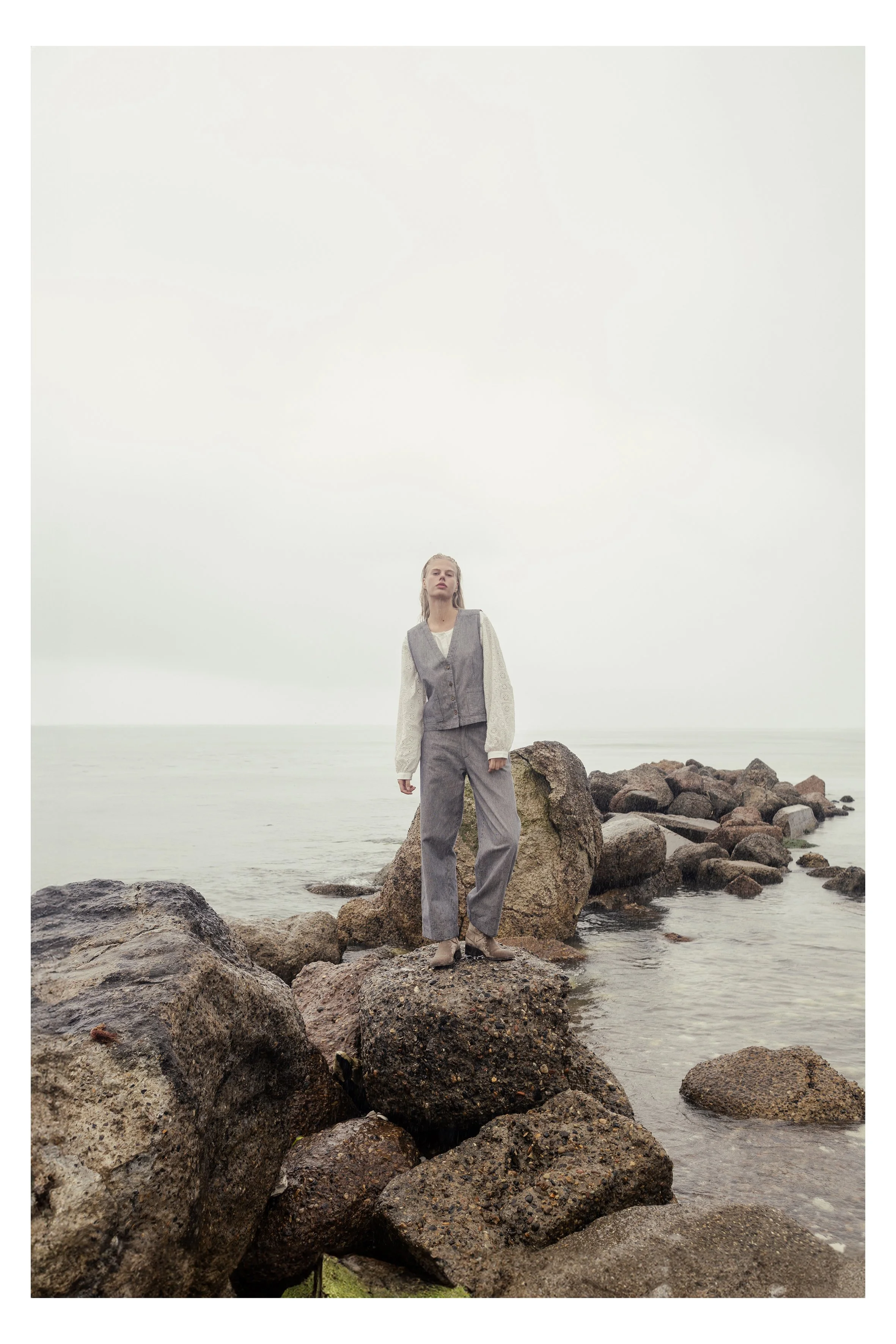 A woman standing on rocks along a shoreline, with water and cloudy sky in the background, dressed in a gray vest and pants, with her hair wet.