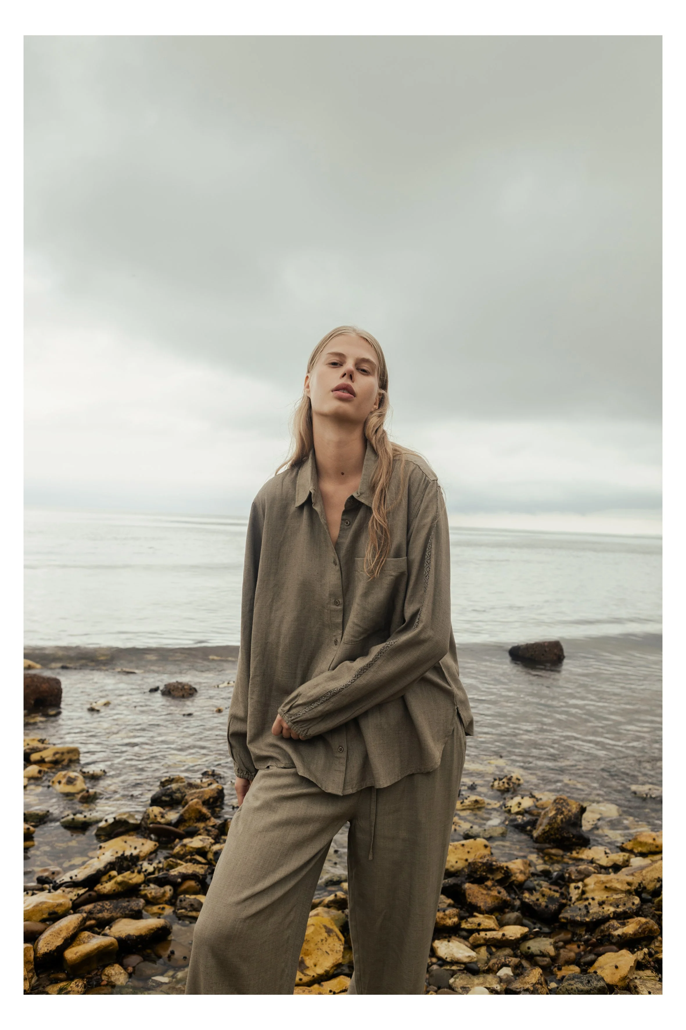 A woman in beige oversized clothing stands on rocky shoreline by the water under cloudy sky.