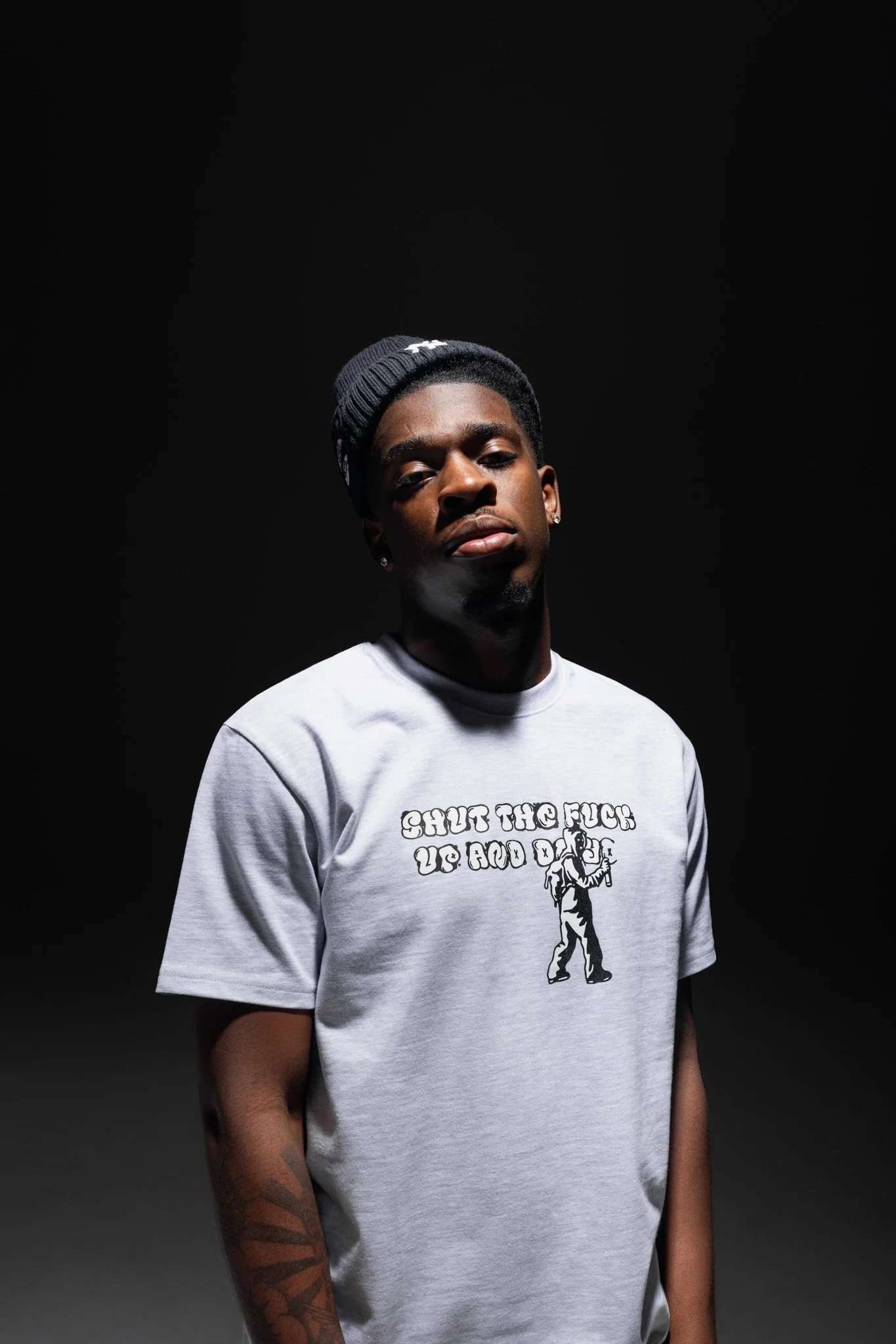 A young man with dark skin and short hair, wearing a black knit beanie and a white t-shirt with a graphic and text that reads "SHUT THE F*** UP AND DANCE." He has earrings and a tattoo on his right arm. The background is black.