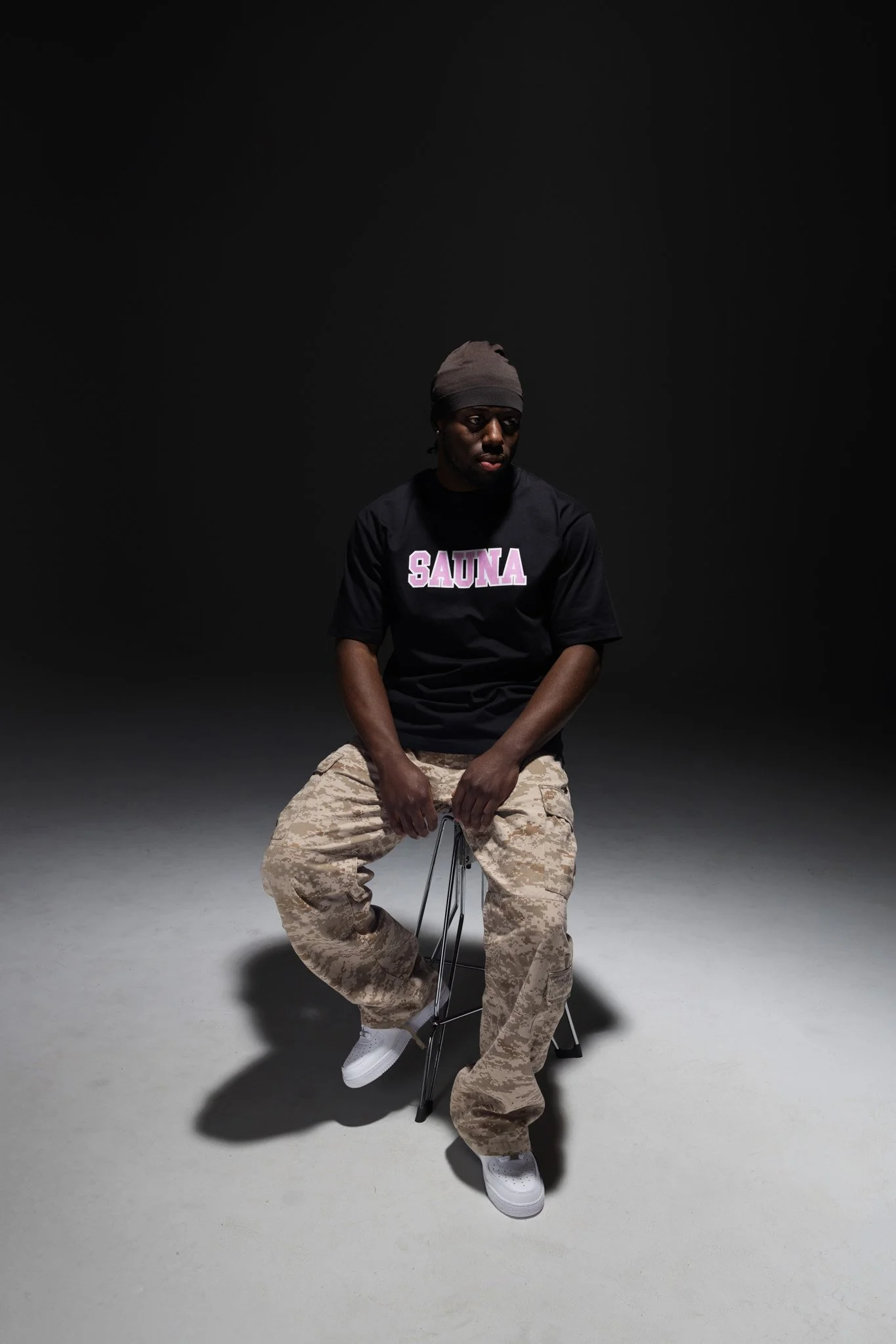 A young man sitting on a stool in a dark studio with a black background, wearing a gray beanie, a black T-shirt with the word "SAUNA" in pink and white letters, beige camouflage cargo pants, and white sneakers.