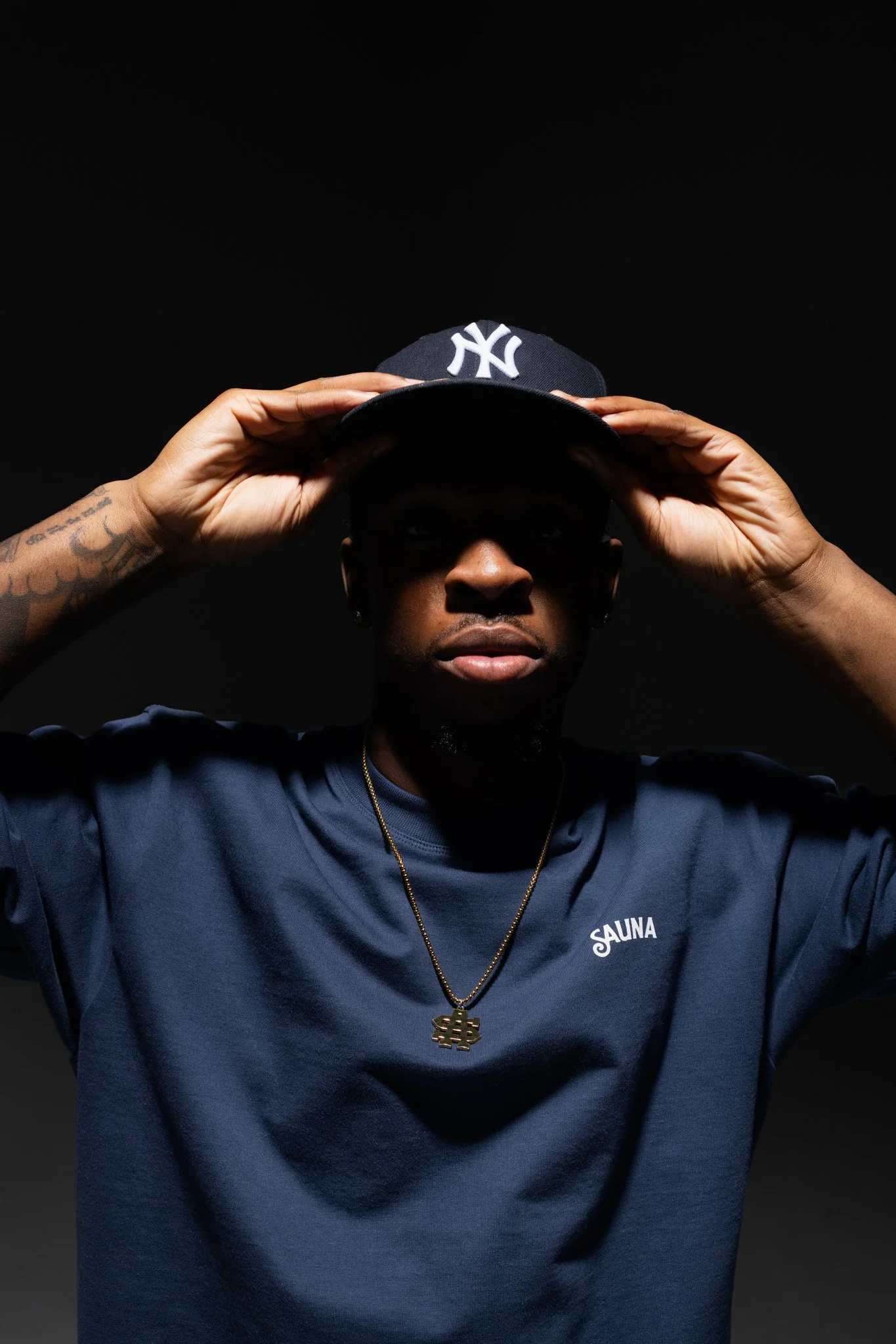 A person wearing a dark blue shirt with the word 'SAUNA' on it and a gold necklace with a dollar sign pendant. They are holding a black New York Yankees cap in front of their face, obscuring their eyes, against a black background.