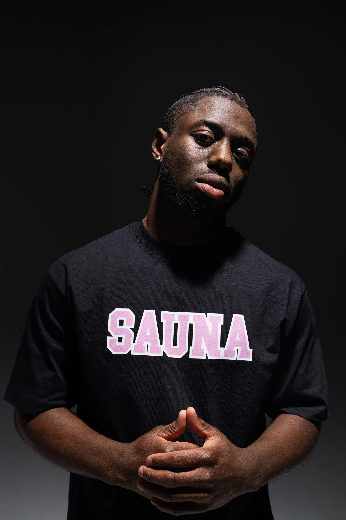A person with dark skin and braided hair, wearing a black sweatshirt with 'SAUNA' written in pink and white, stands against a dark background, with hands clasped in front.