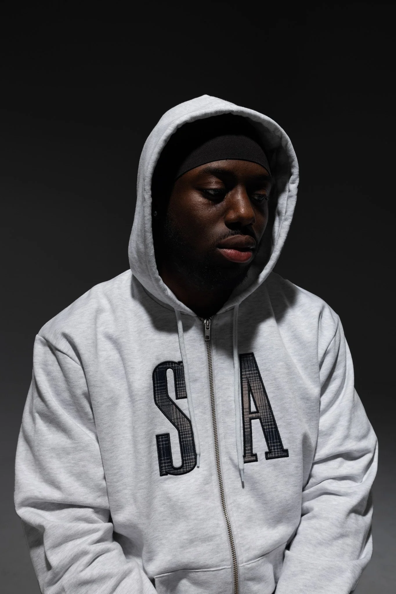 A young man with dark skin, wearing a light gray hoodie with the words 'USA' on the front, and a black head covering, looking down with a serious expression.