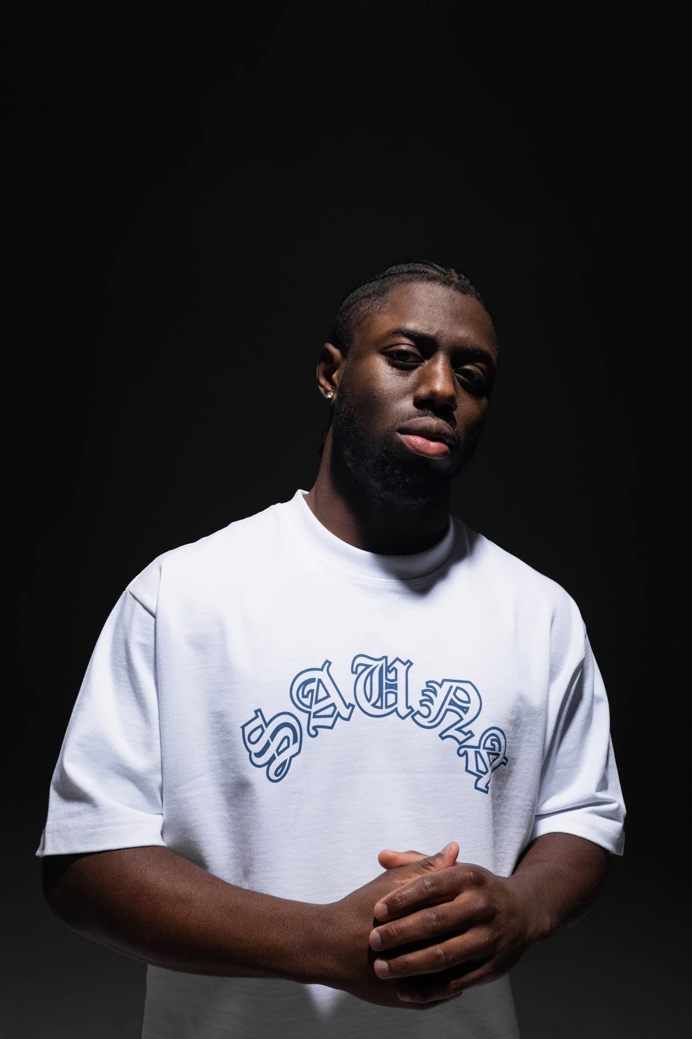 A young man with braided hair and earrings wearing a white sweatshirt with a blue design, standing against a dark background.