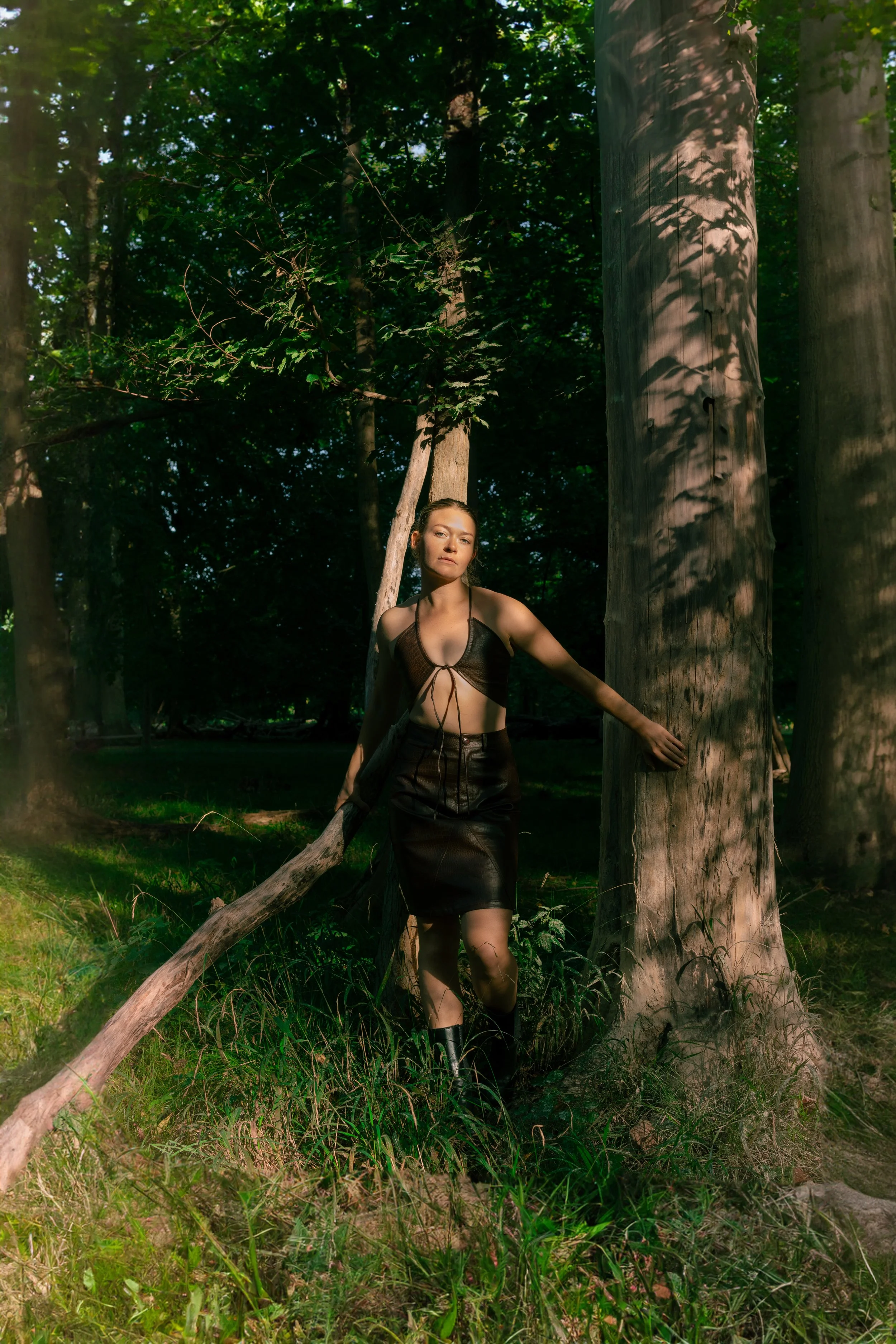 A young woman with brown hair and a leather outfit standing among trees and tall grass in a forested area with sunlight creating shadows on the trees.