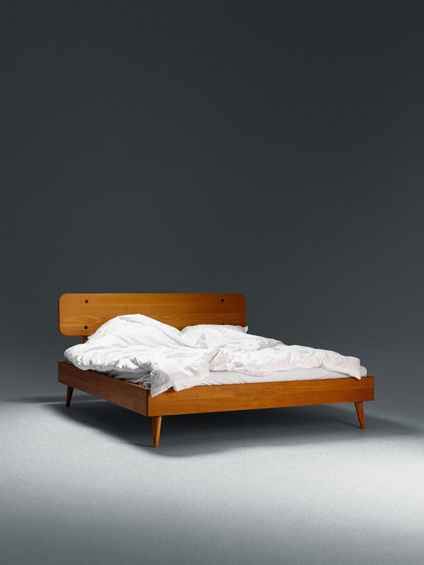 A wooden bed frame with a white, unmade bedsheet and pillows against a dark gray wall background.