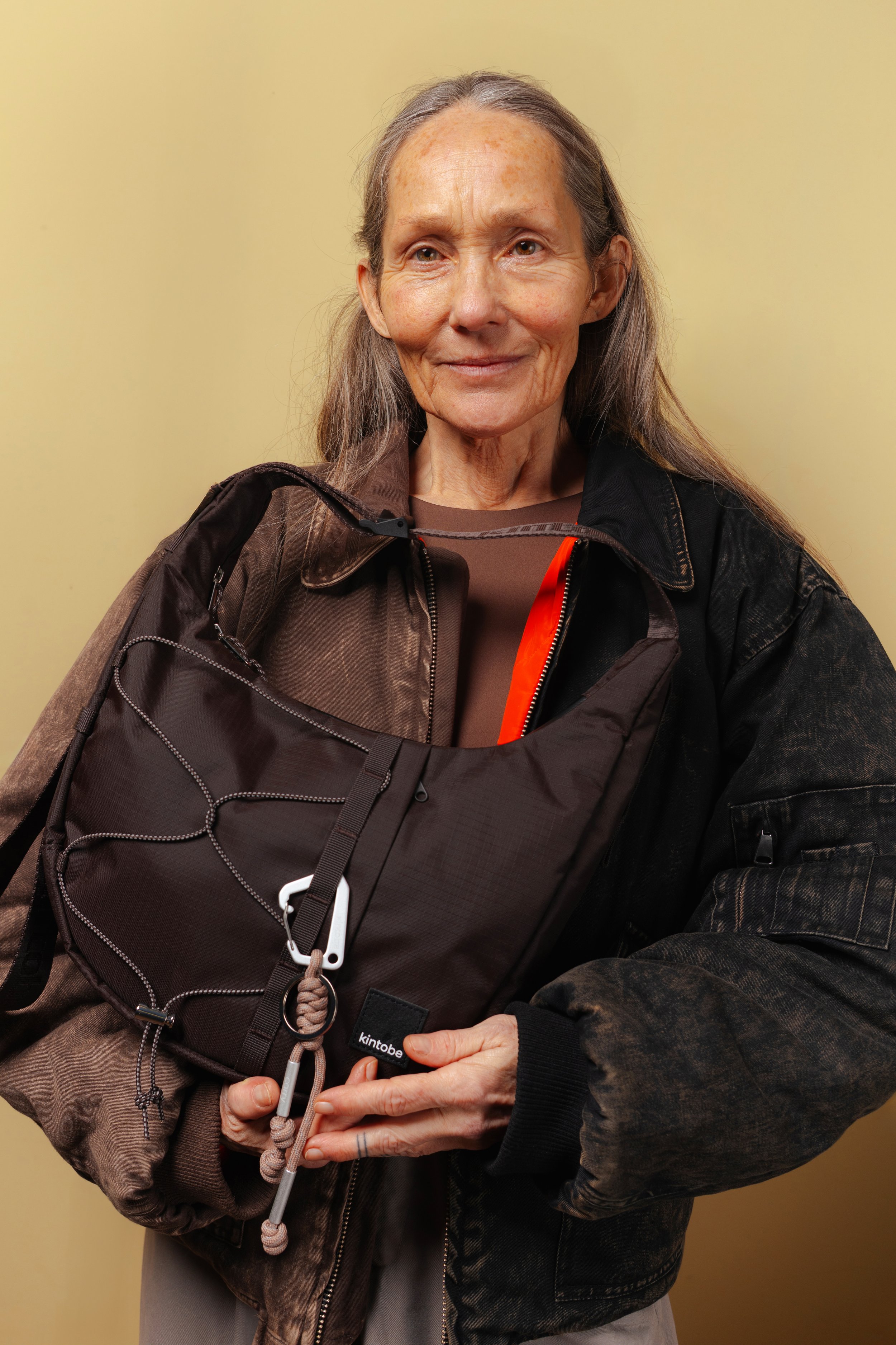 A woman with long hair and freckles wearing a brown shirt, black jacket, and a brown backpack with a carabiner and rope, standing against a plain yellow wall.