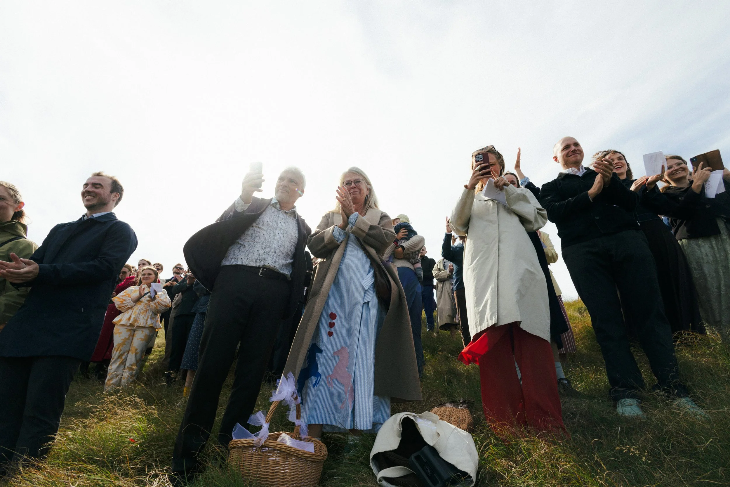 En gruppe mennesker står på en græsmark og ser op mod himlen, nogle tager billeder eller videoer med deres telefoner. De ser glade og engagerede ud.