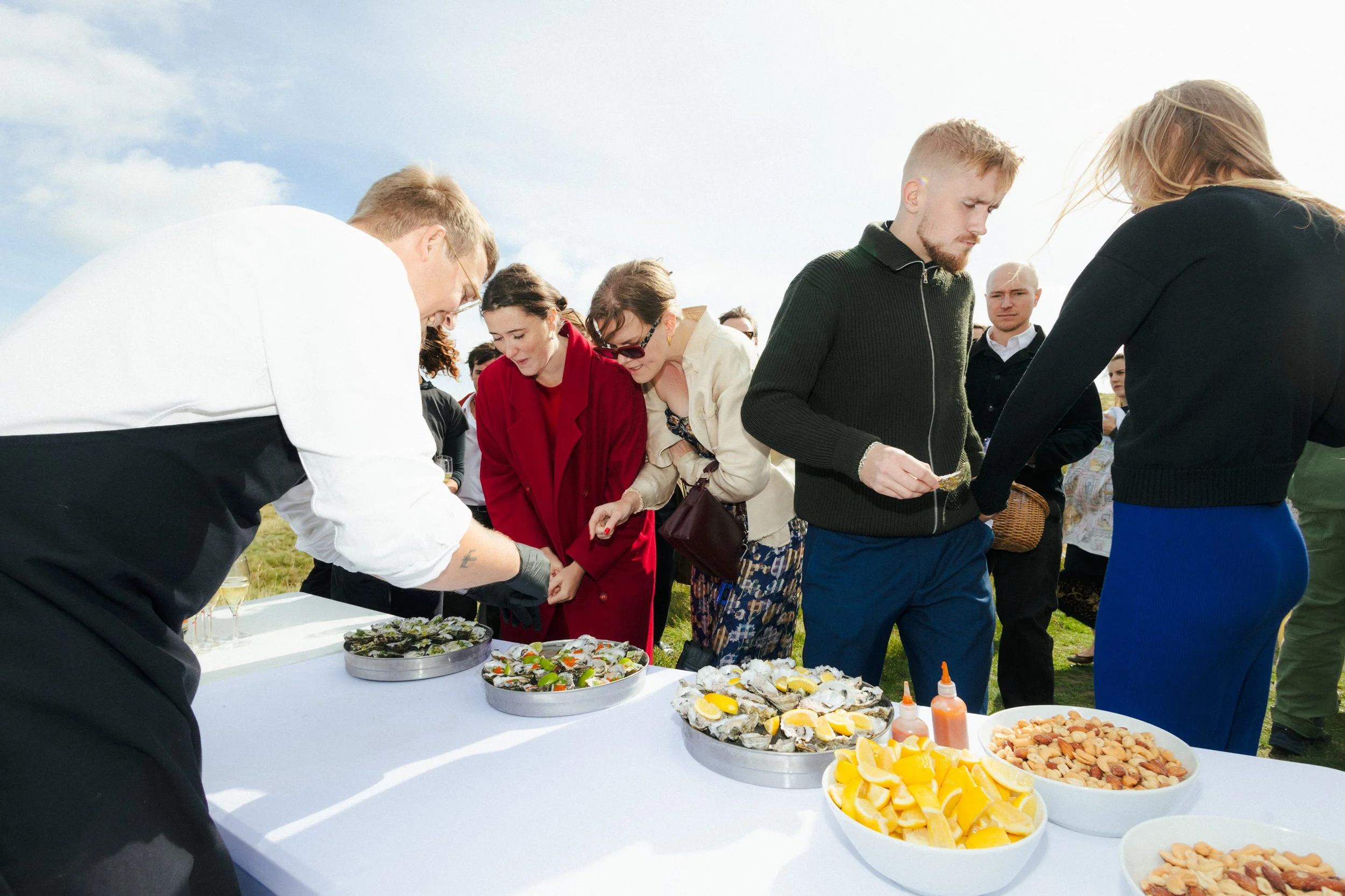 Folk, der står i kø foran en buffet med forskellige retter ved en udendørs begivenhed under en klar himmel.