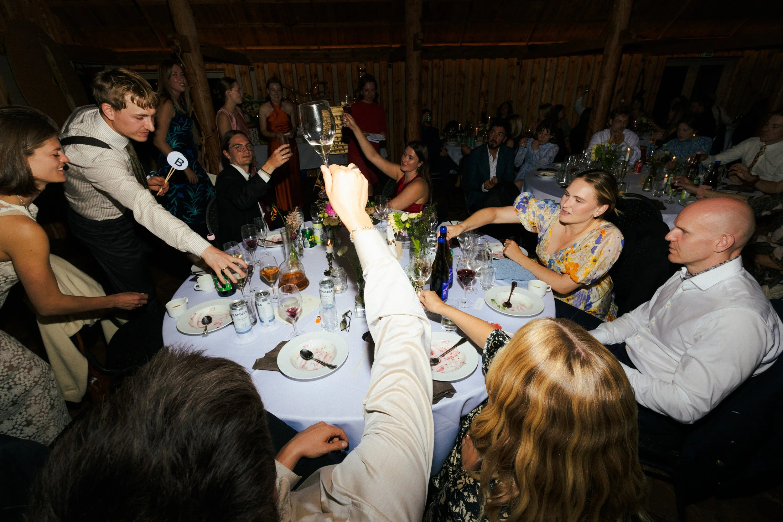 En gruppe mennesker sidder rundt om et bord til en festlig begivenhed, hvor nogle skåler med champagne hæves, mens andre spiser og taler. Der er blomsterdekorationer og tomme tallerkener på bordet.