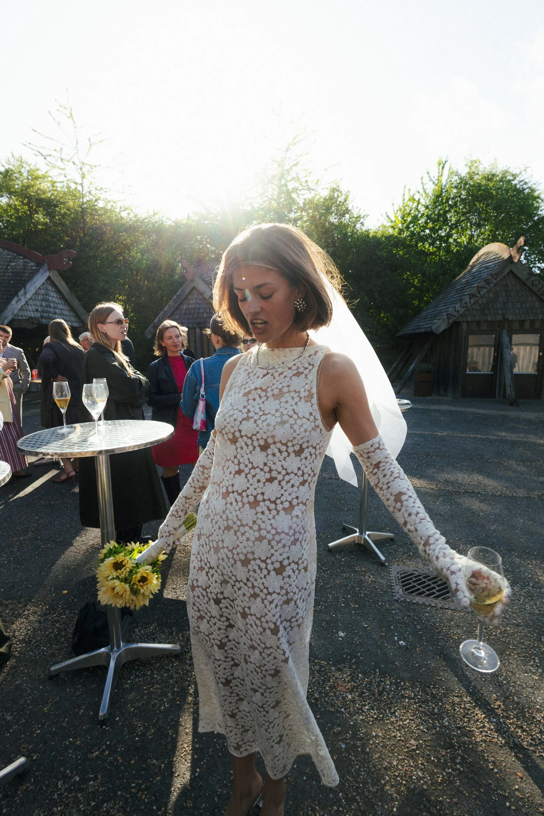 En kvinde i en lang, hvid blondekjole holder en blomsterbukett og står ved en bar udendørs sammen med flere mennesker, der taler og drikker, med grønne træer i baggrunden og solen, der skinner.